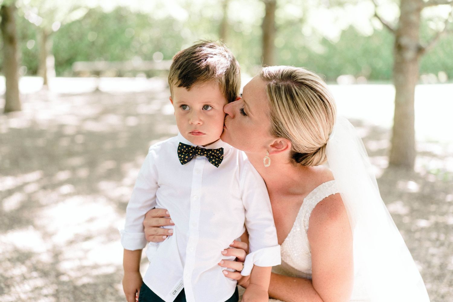 Sweet wedding day moment in sunlit outdoor setting with formal attire and bow tie.