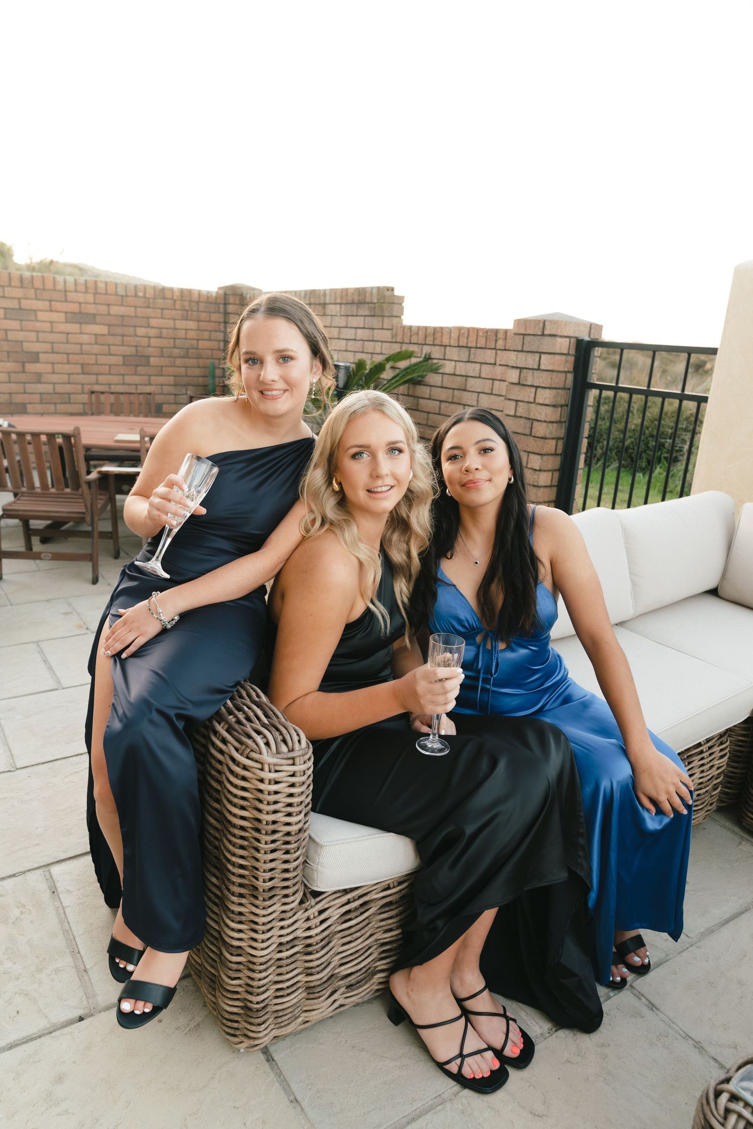Three friends in formal dresses pose together on wicker patio furniture during an evening celebration.