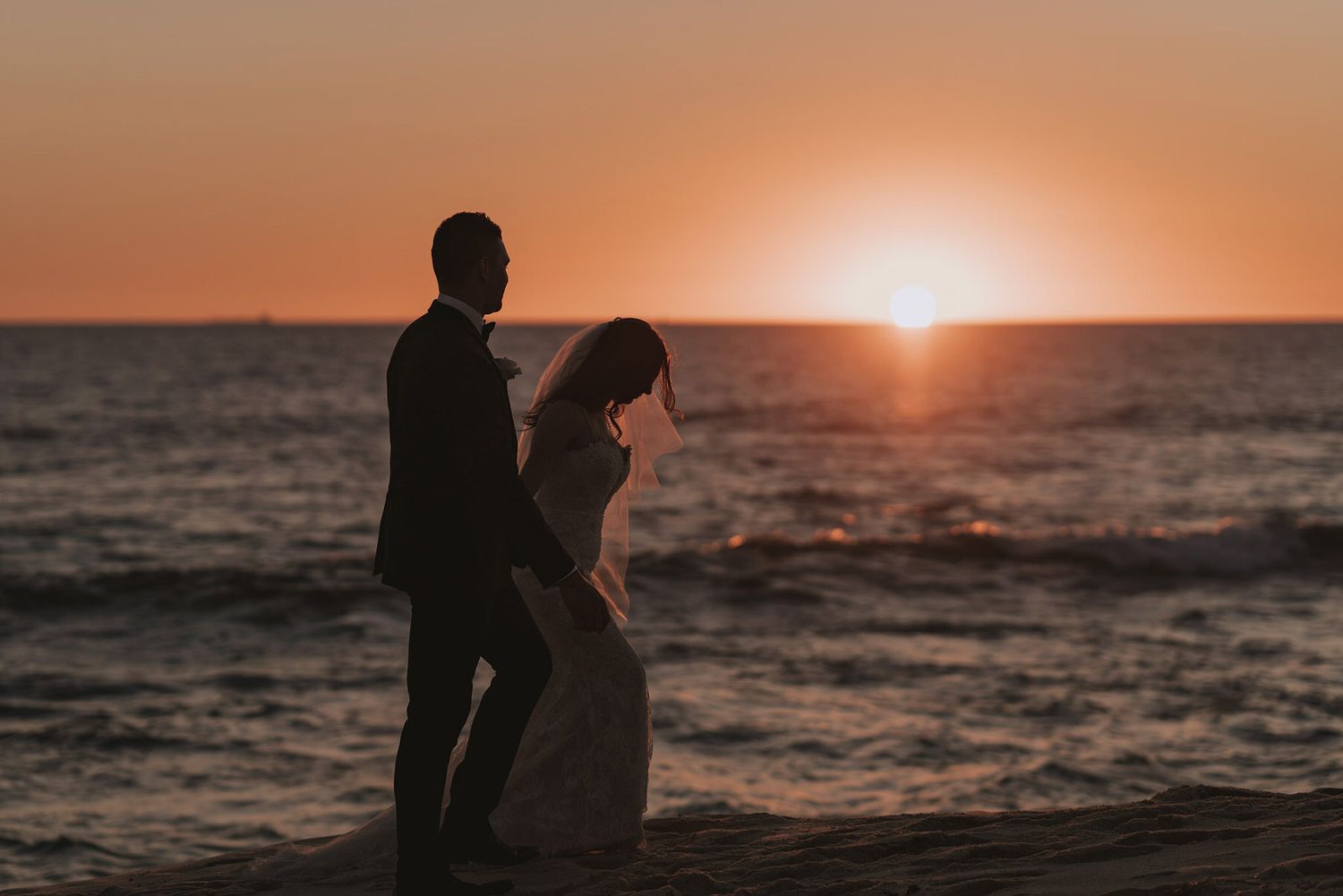 Two silhouettes standing together at sunset on a beach with waves crashing and an orange sky.