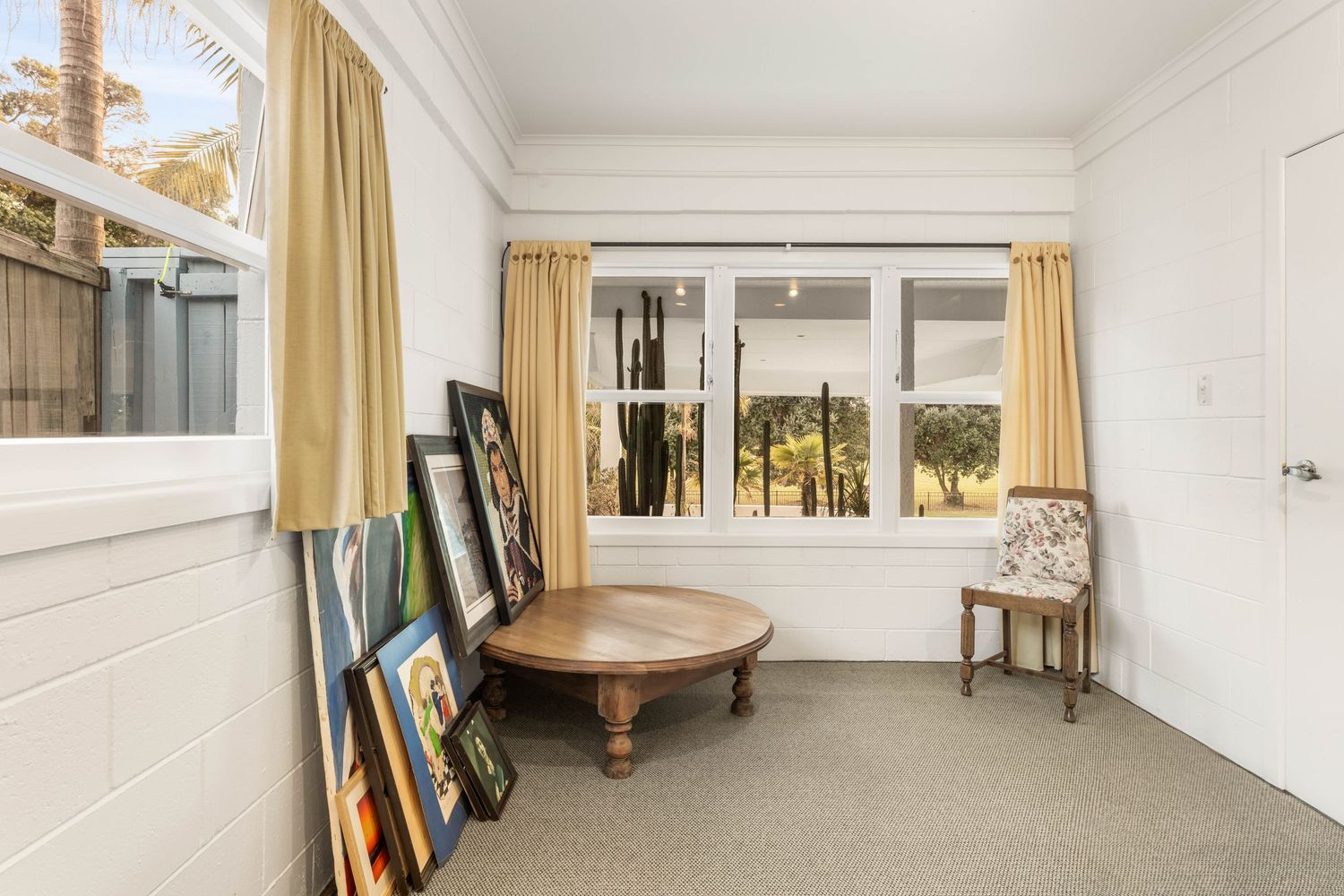 Bright room with round wooden table, yellow curtains, and artwork leaning against white walls.