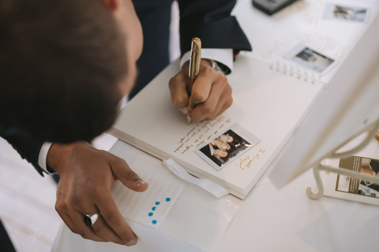 A person's hand writes in a notebook while multiple papers and documents are spread across a white desk surface.