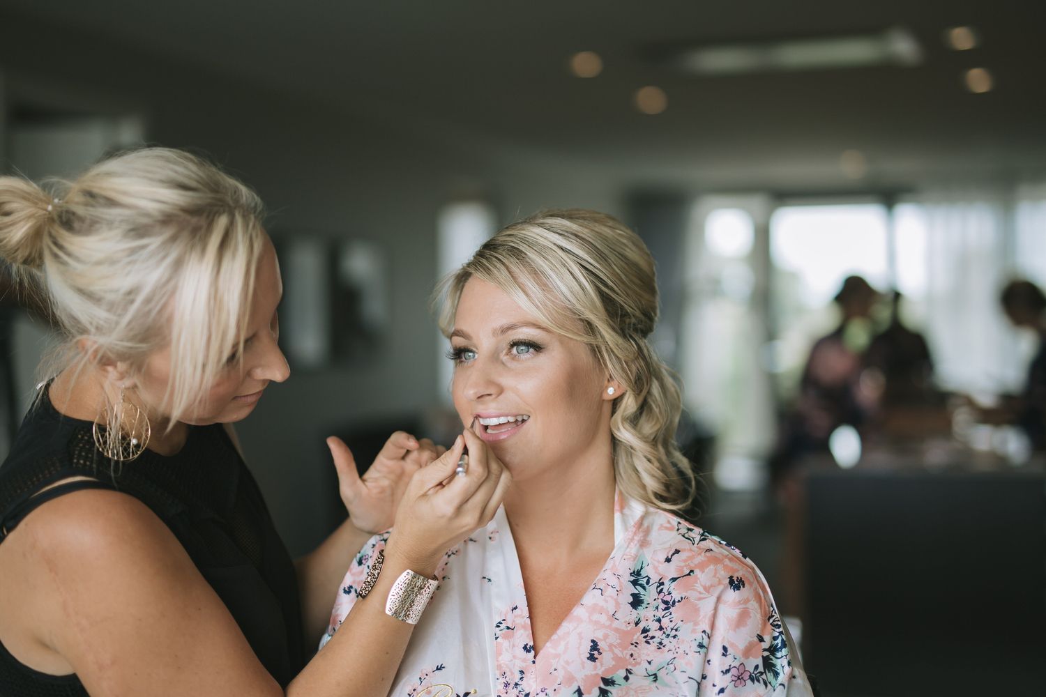 Two women share a happy moment during wedding day preparations as makeup is being applied in a light-filled room.