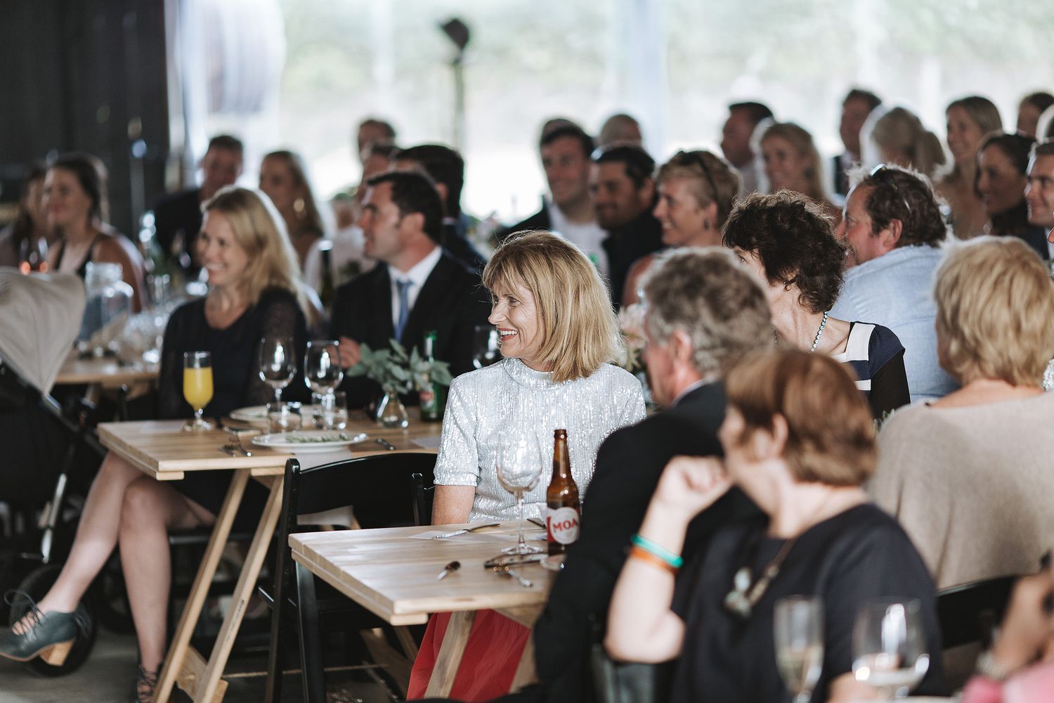 People sit at wooden tables for a dining event in a venue with large windows and natural lighting.