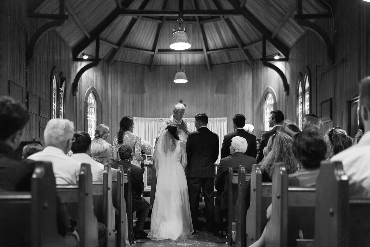 A black and white photo of a wedding ceremony in a traditional church sanctuary with wooden pews and arched windows.