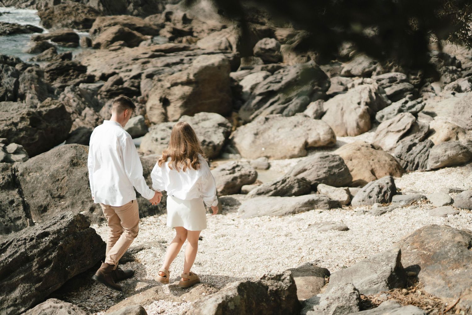 Young couple holding hands while walking along rocky coastal shoreline in white summery outfits.