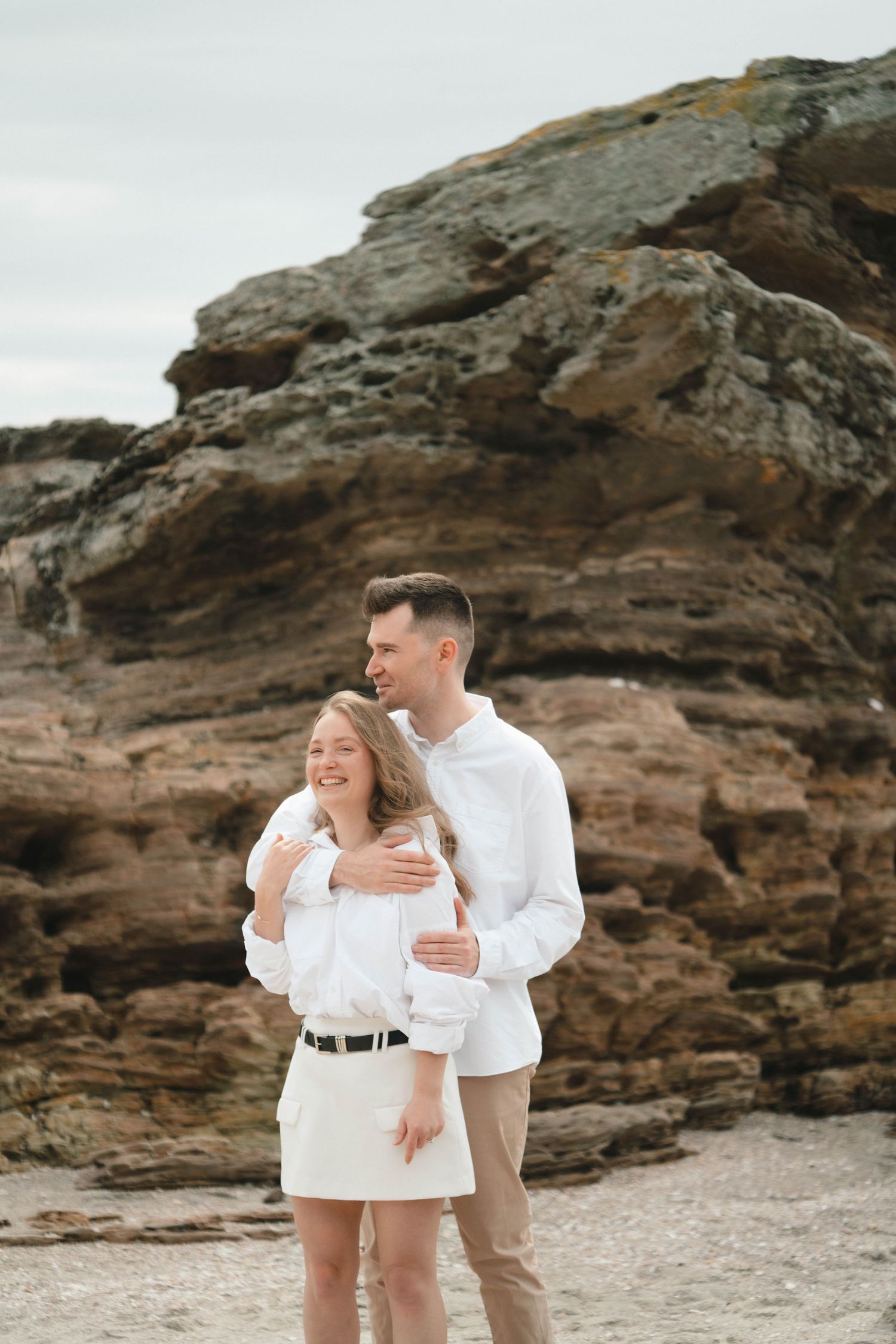 A couple laughs together while embracing on a rocky beach wearing matching white tops.