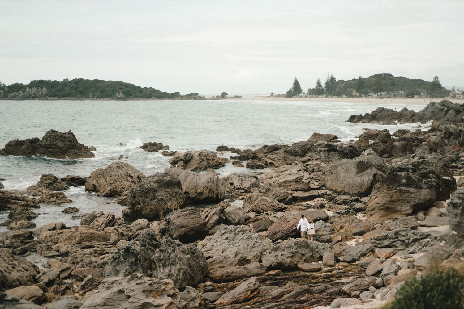 Rocky coastal shoreline stretches along the beach with waves crashing against rugged outcrops and scattered boulders at low tide.