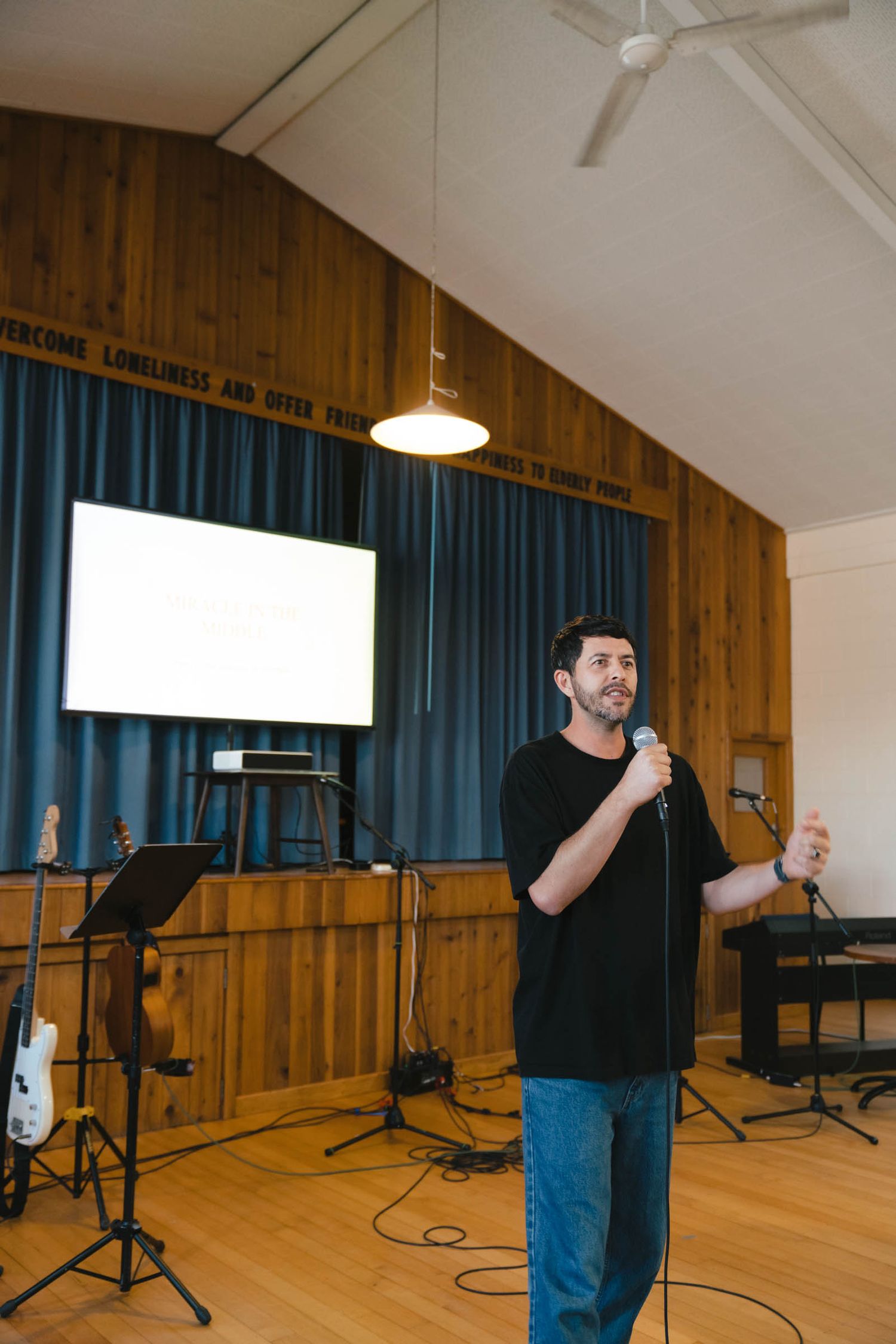 A speaker gestures while presenting in a performance space with wood paneling and stage lighting.