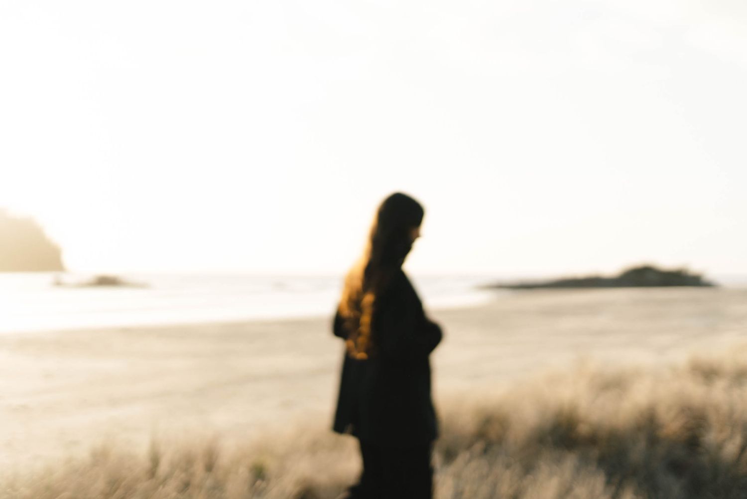 Blurred silhouette figure stands against warm sunset sky on coastal dunes.