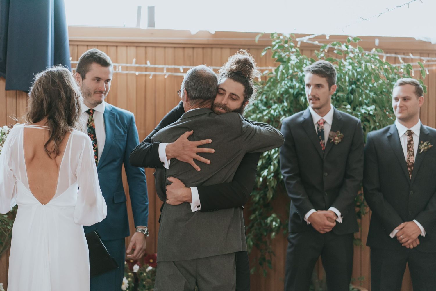 A wedding party lineup stands in formal attire against a wooden paneled wall at an indoor venue.