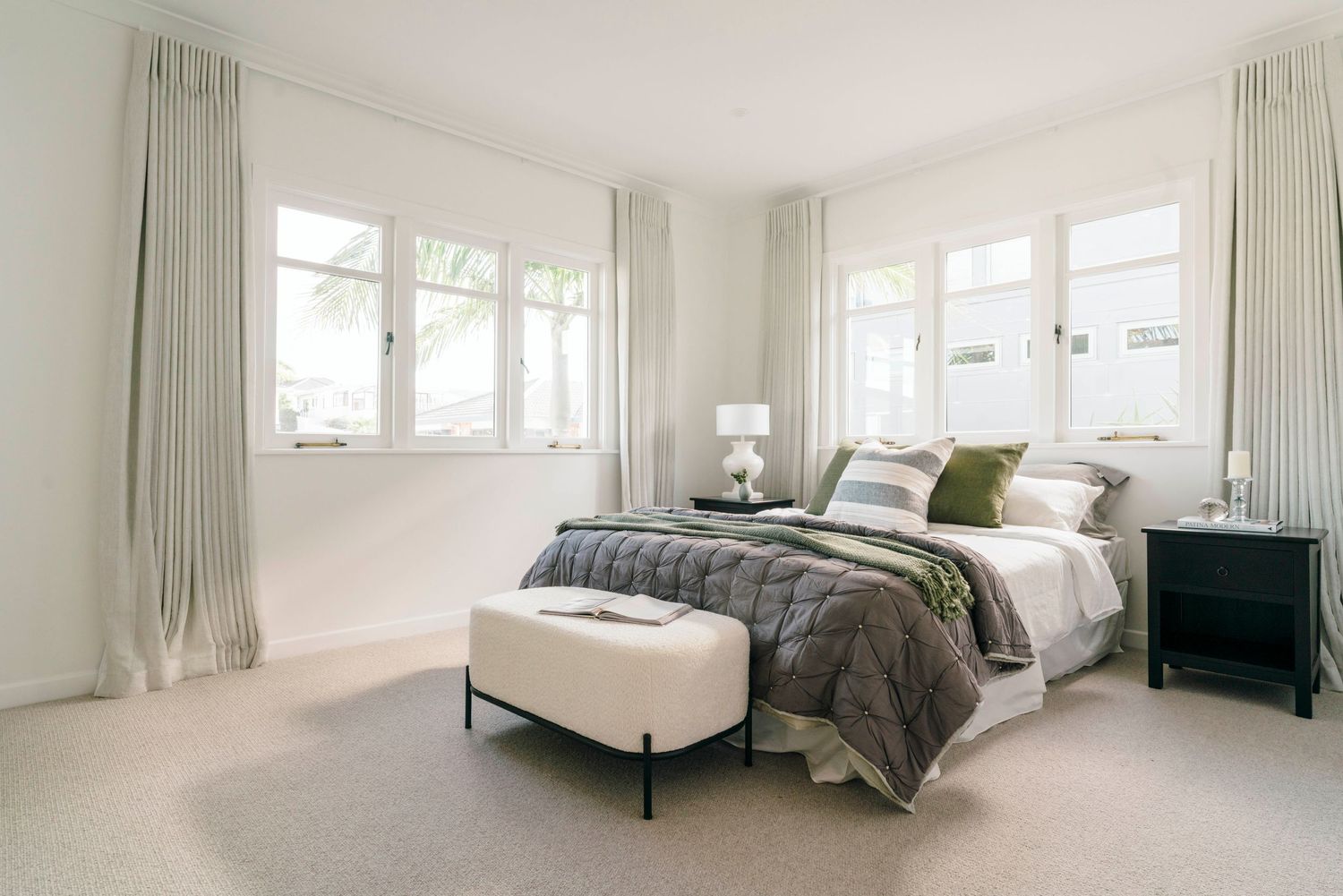 Serene white bedroom with large windows, gray bedding, and upholstered bench at foot of bed.