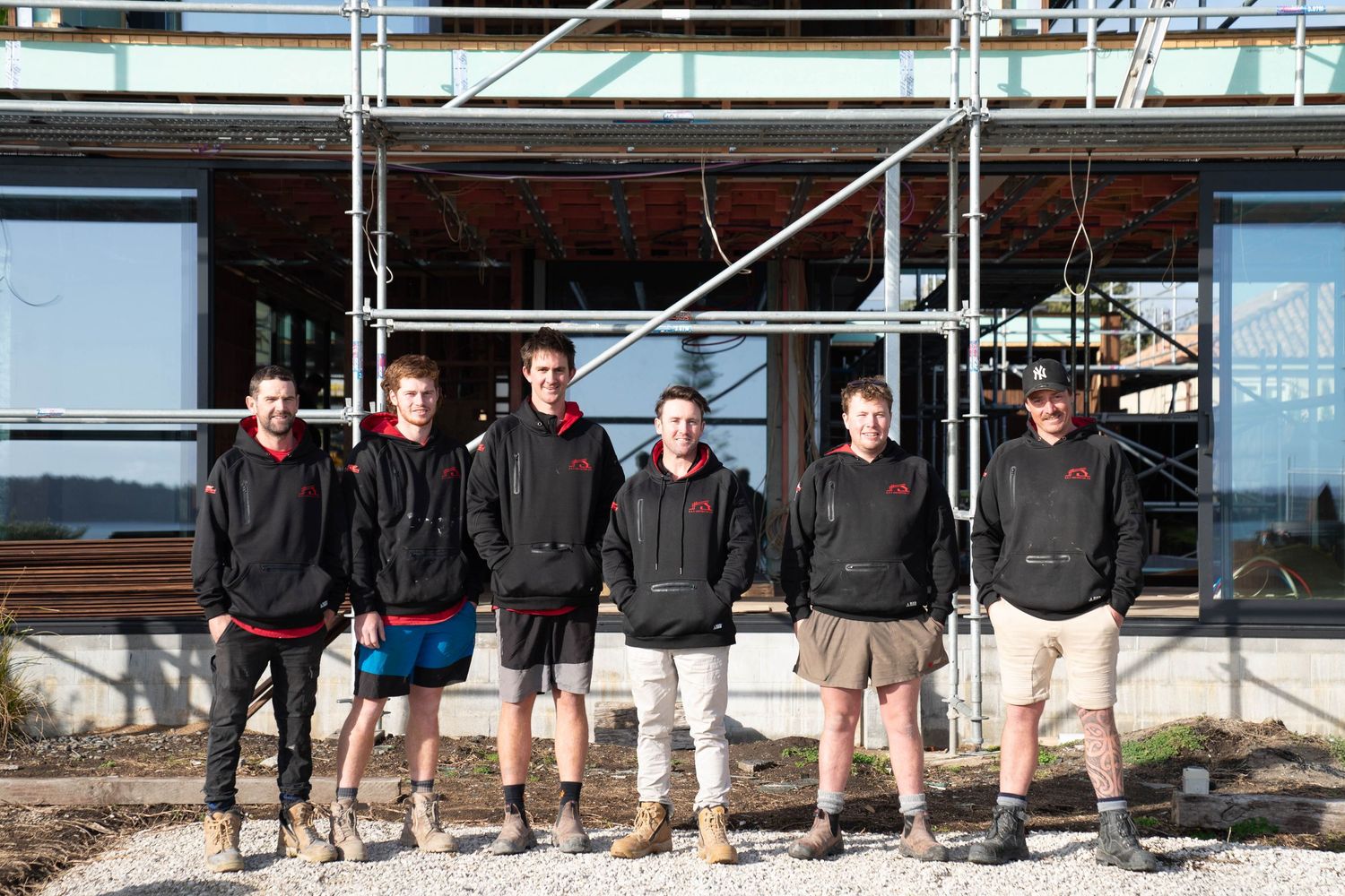 Group of construction workers in black jackets and shorts standing together in front of scaffolding.