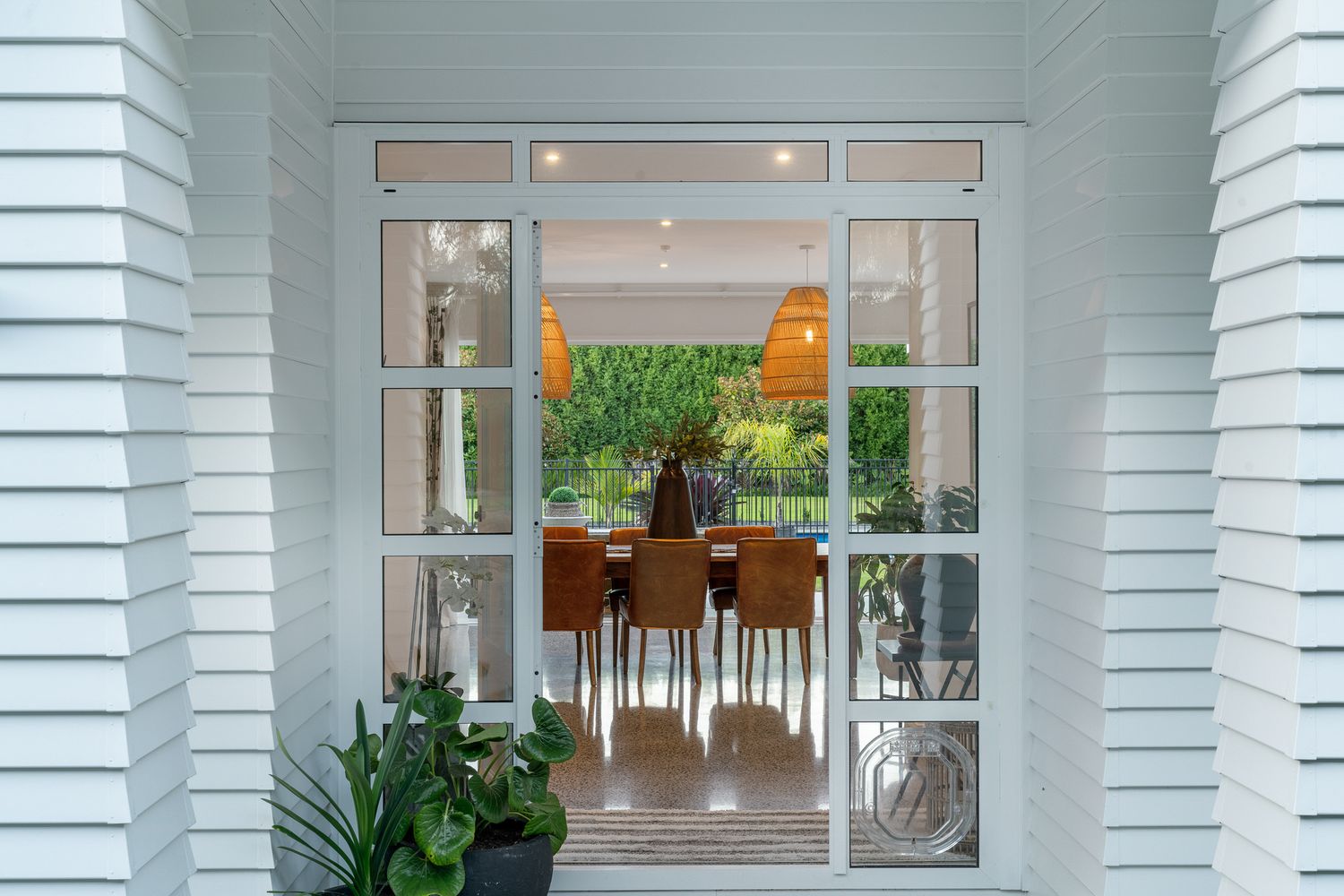 View through French doors showing an elegant dining area with rattan pendant lights and wooden furniture.