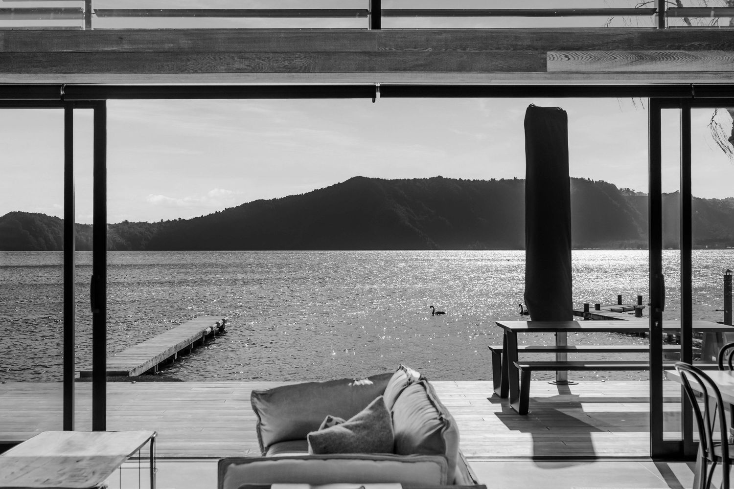 Black and white view of a lakeside deck with lounger chairs overlooking a tranquil water scene and distant hills.