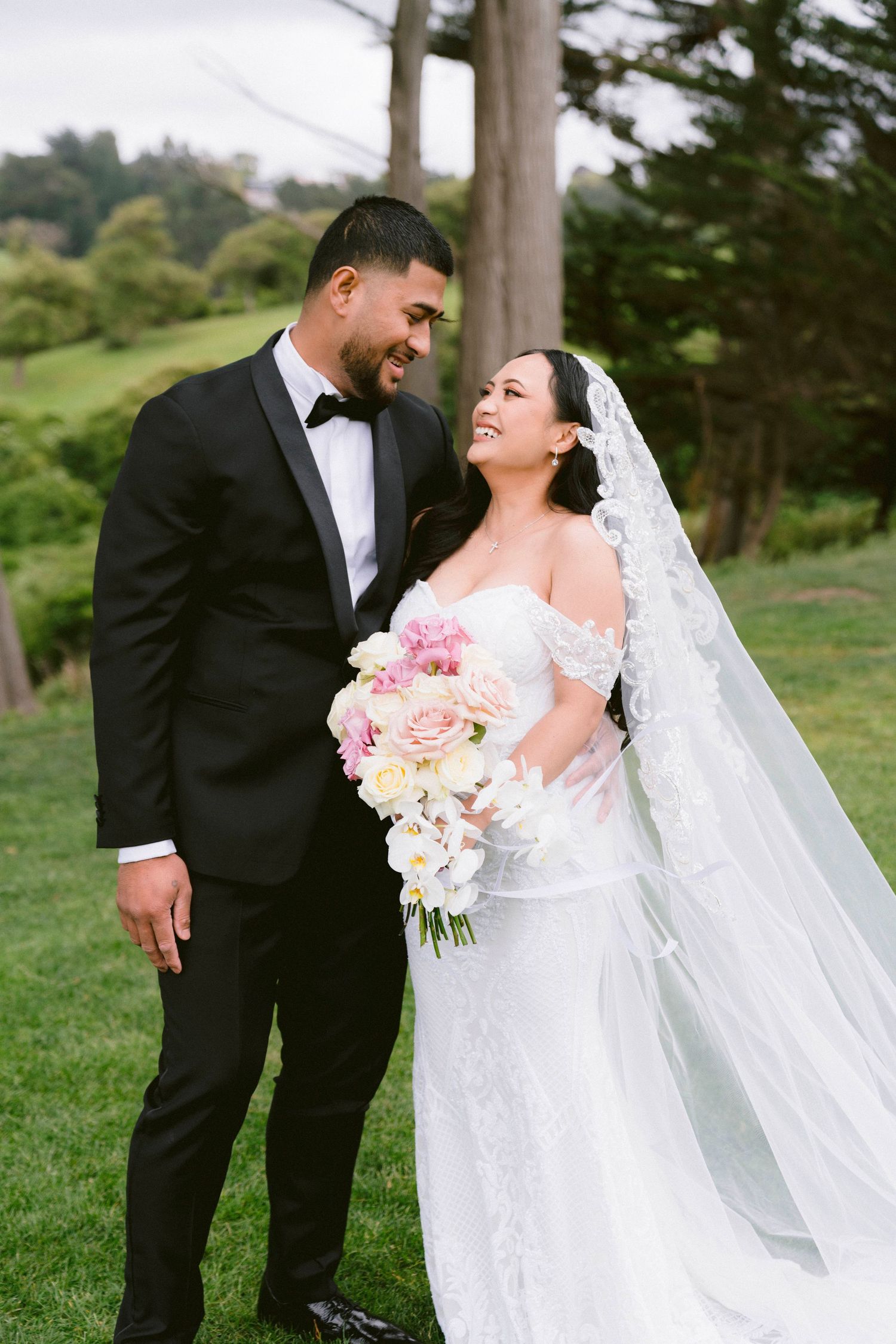 Wedding couple shares a joyful moment outdoors with a bouquet of pink and white flowers.