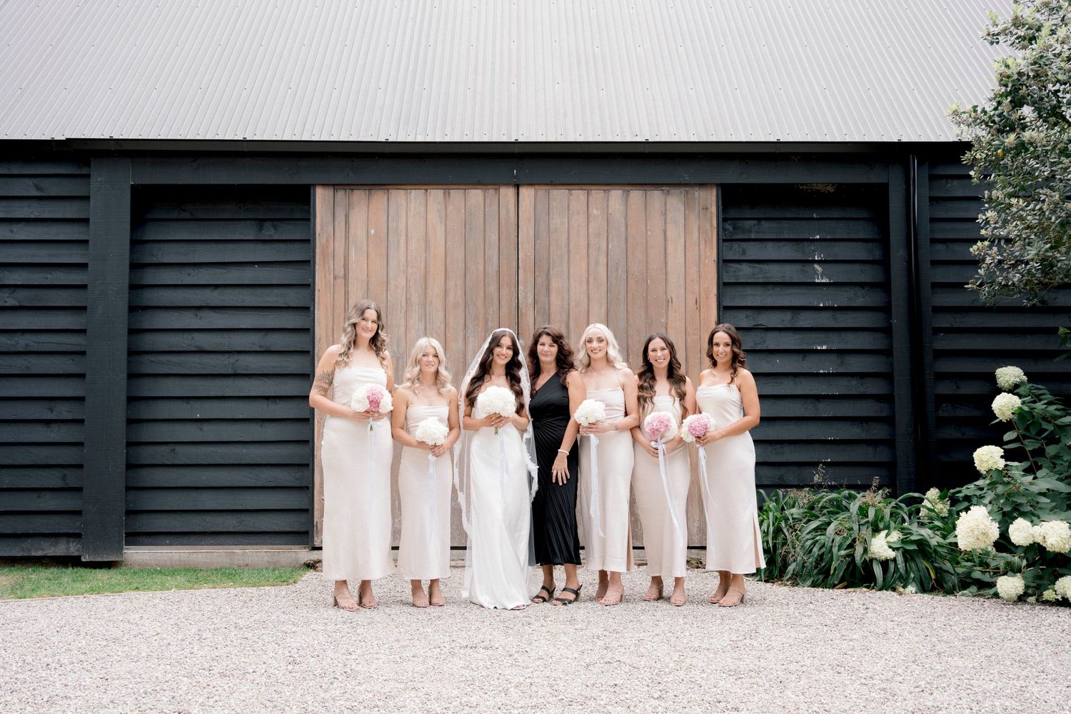 Bridesmaids in light cream dresses pose in front of a modern black barn building.