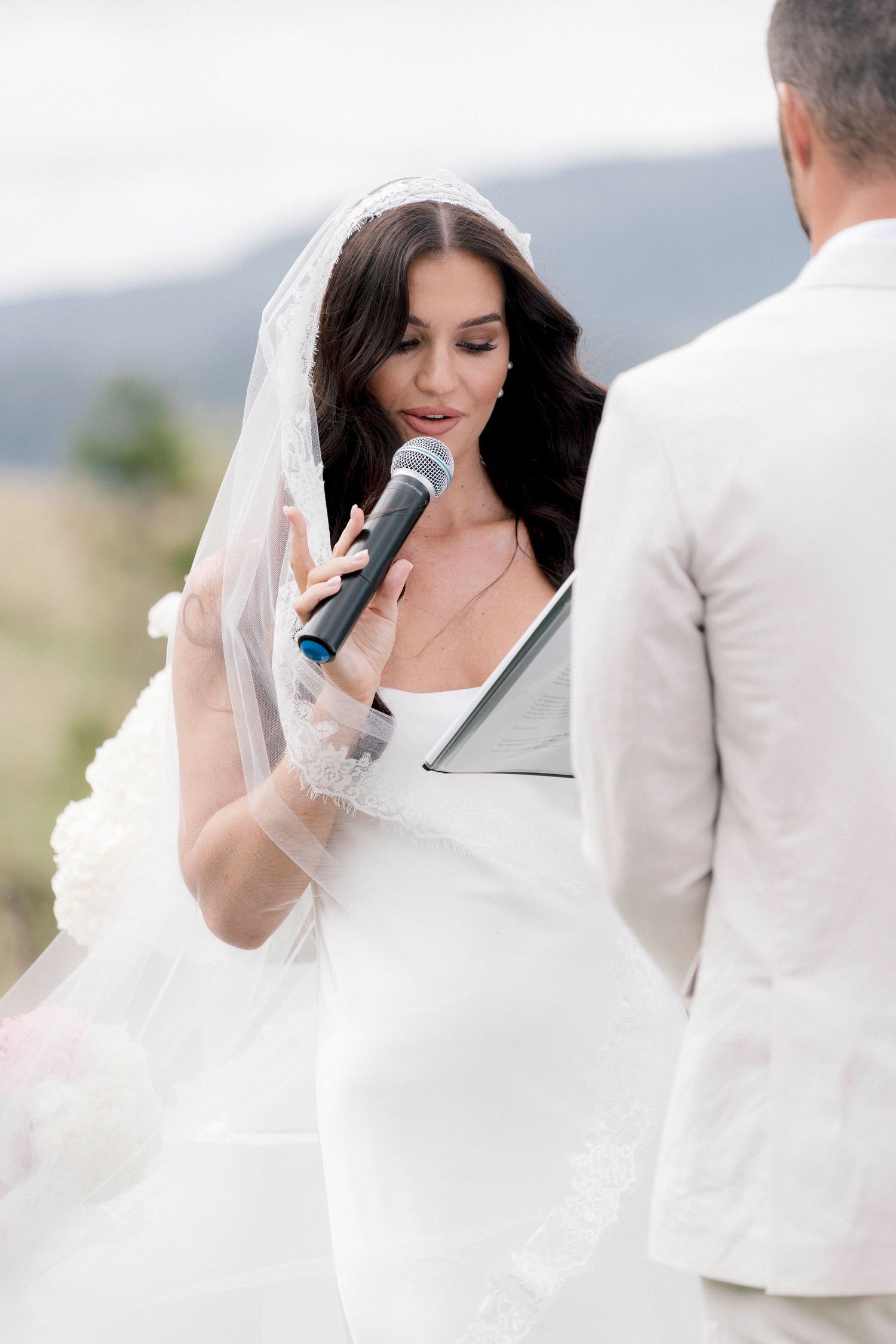Bride wearing lace veil recites vows with microphone during scenic mountaintop wedding ceremony.