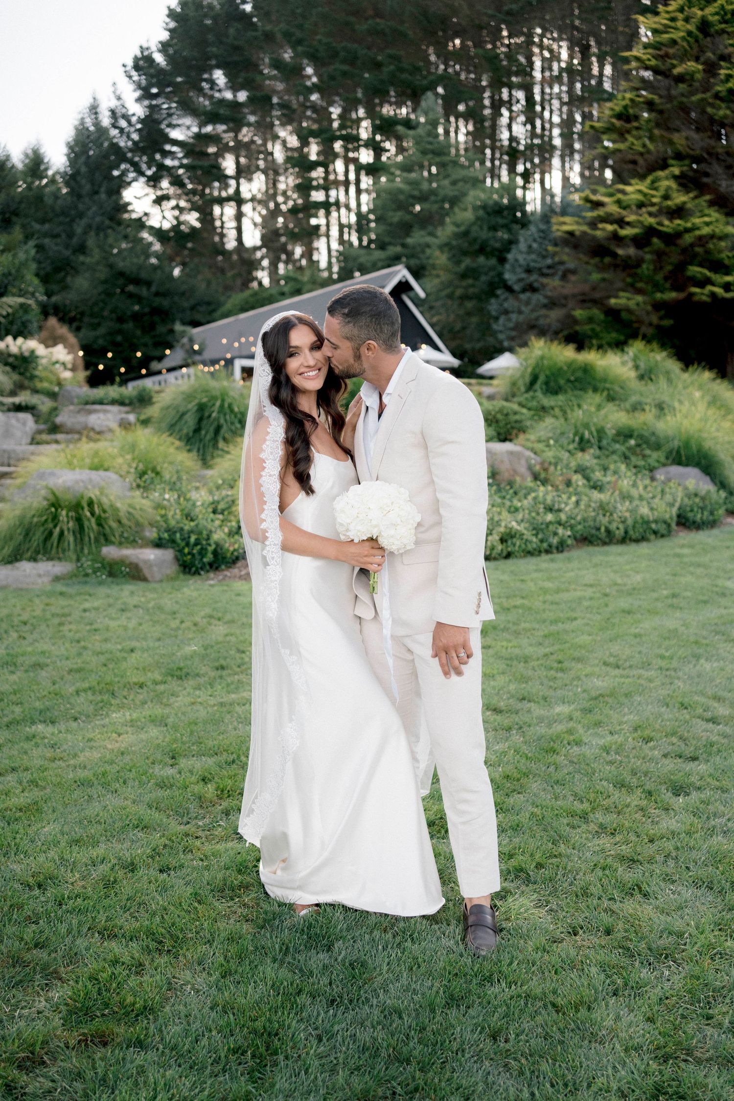 Romantic wedding couple in white embracing on landscaped hillside with evergreen forest backdrop.
