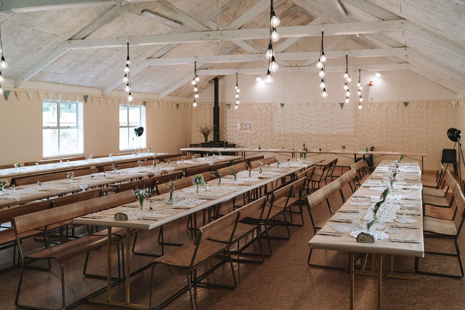 Long wooden folding tables and chairs arranged in rows inside a bright rustic hall with exposed ceiling beams and string lights.
