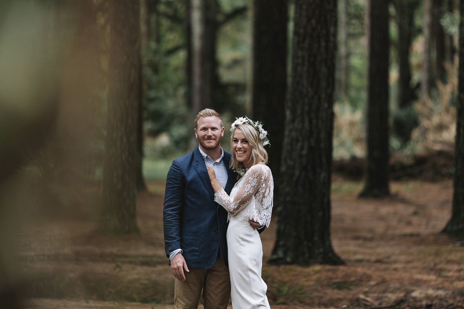 Joyful couple embraces while standing in a forest of tall pine trees on their wedding day.