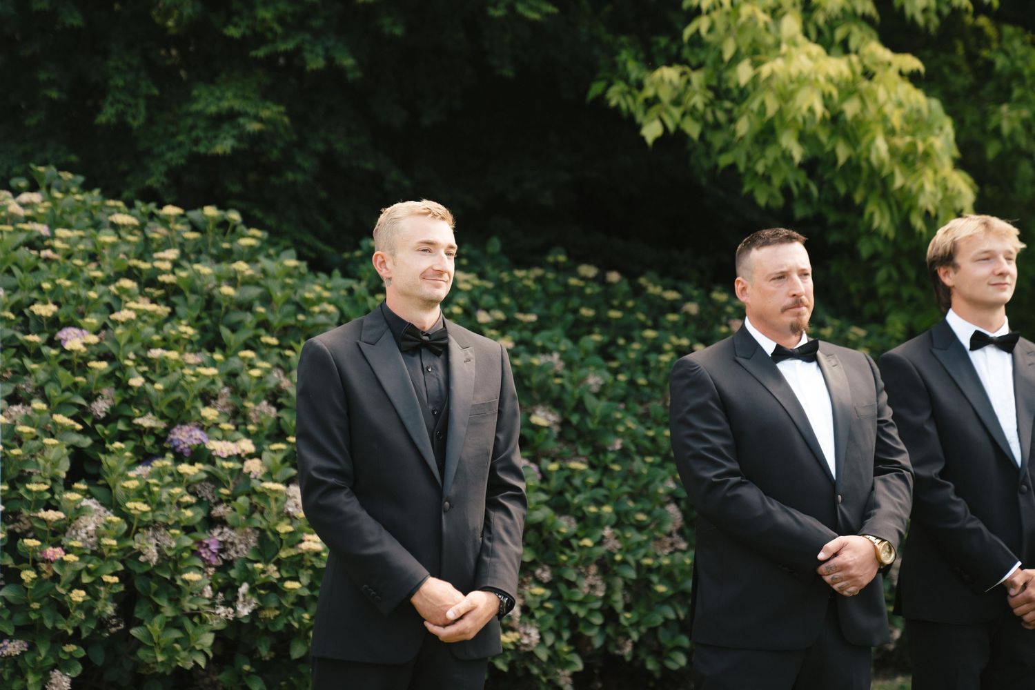 Three groomsmen in formal black attire stand outdoors in front of green foliage and pink flowers during a wedding ceremony.