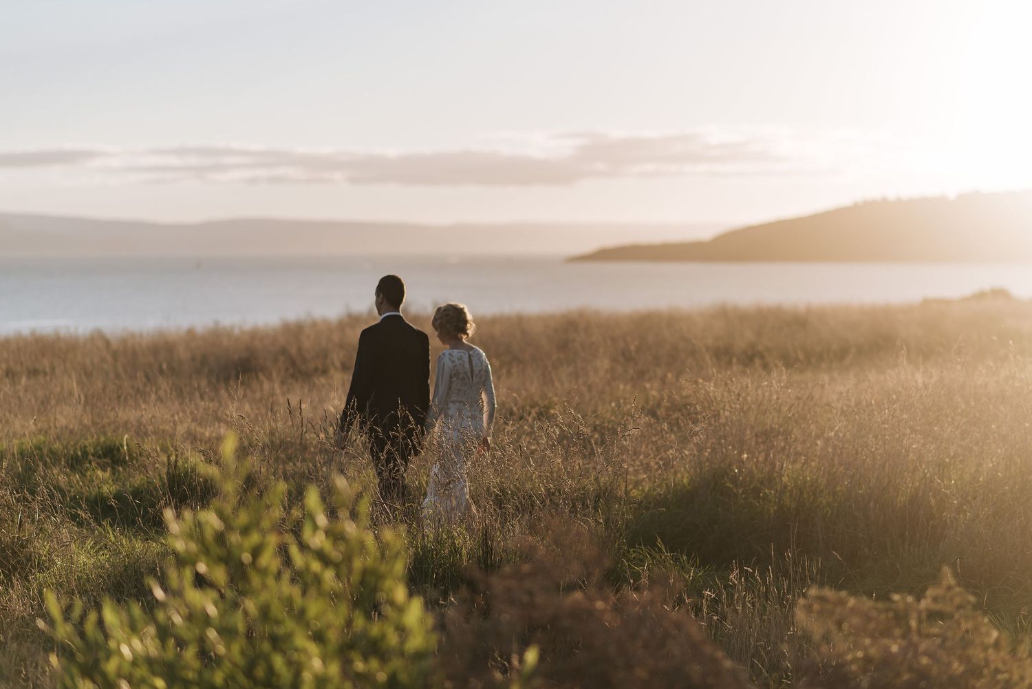 Two people walking through a misty golden field at sunset with hills in the background.