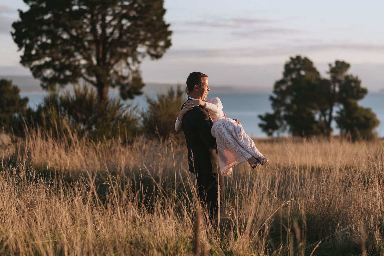 A couple share a romantic moment in tall grass with trees and ocean view in the background.