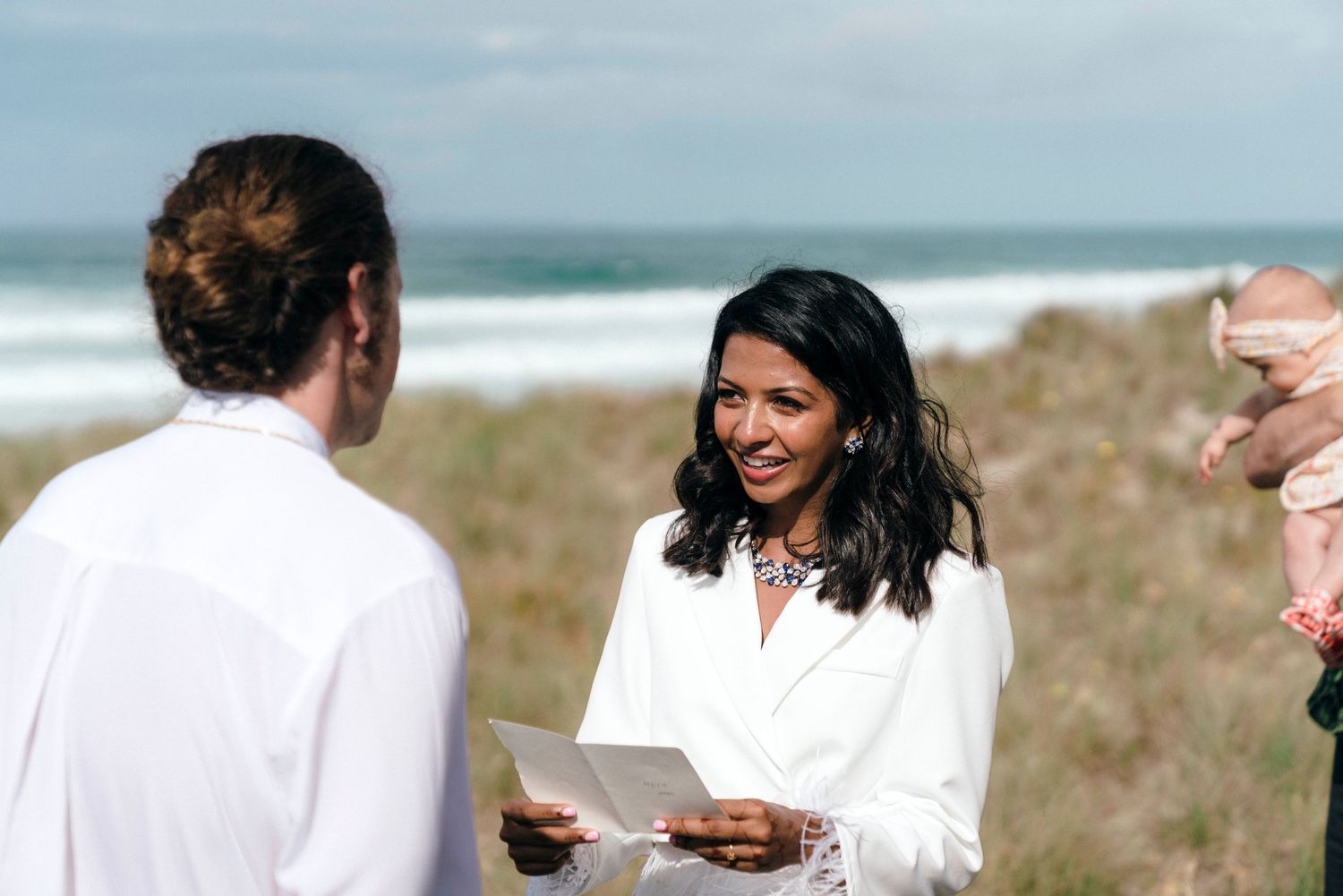 An intimate beach wedding ceremony with attendees gathered on coastal dunes near the ocean.