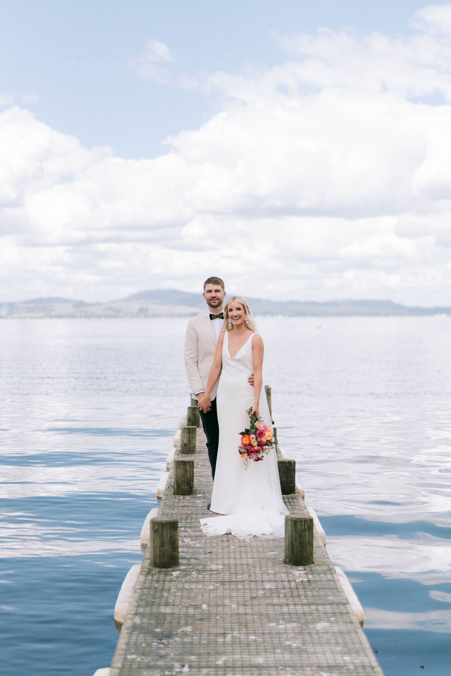 A wedding couple stands on a wooden pier overlooking a serene lake with mountains in the background.