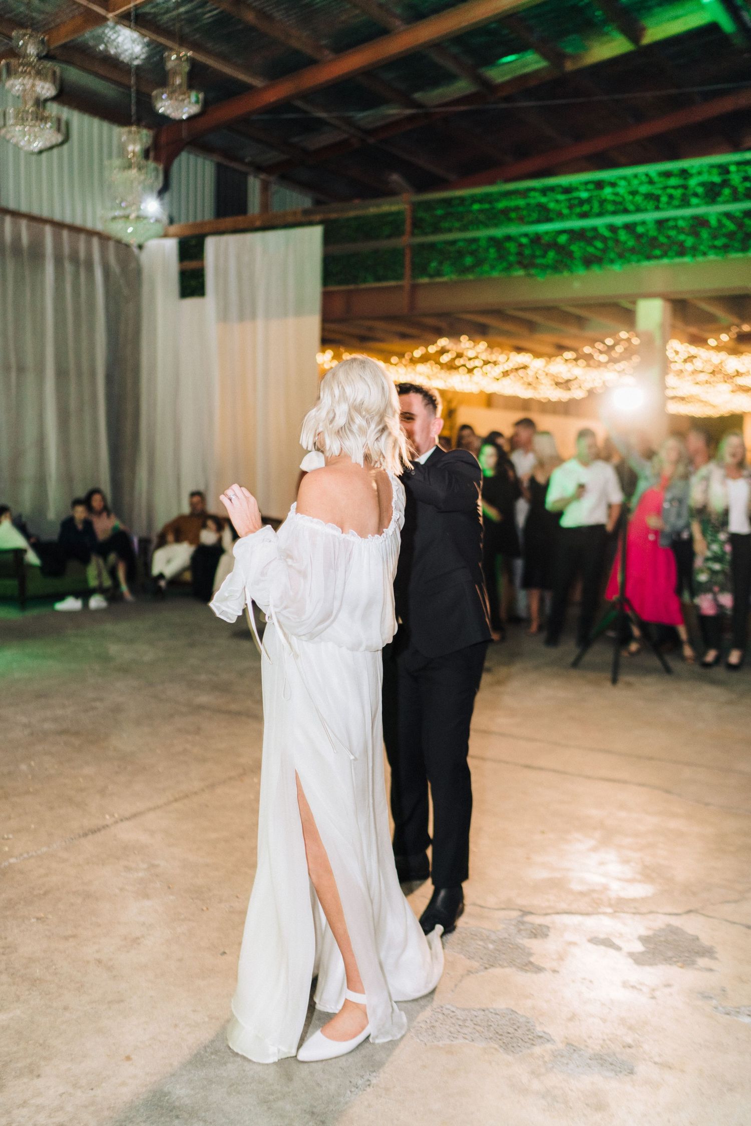 Couples dance on a spacious reception floor under green lighting and string lights.