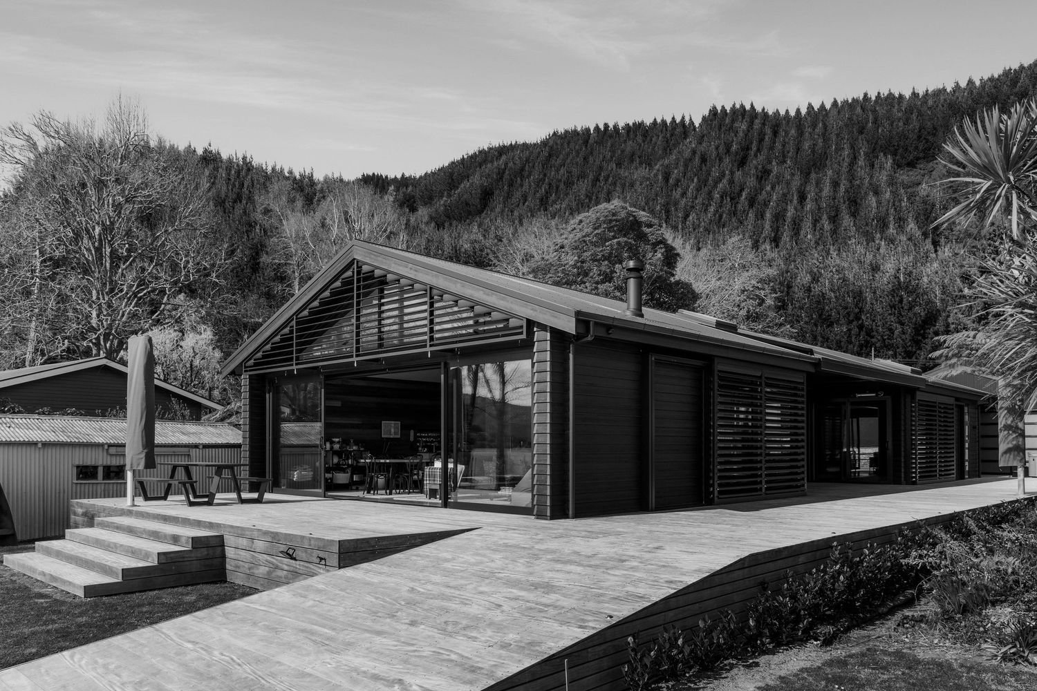 Black and white photo of a modern dark-clad barn-style home with large glazing, set against forested hills.