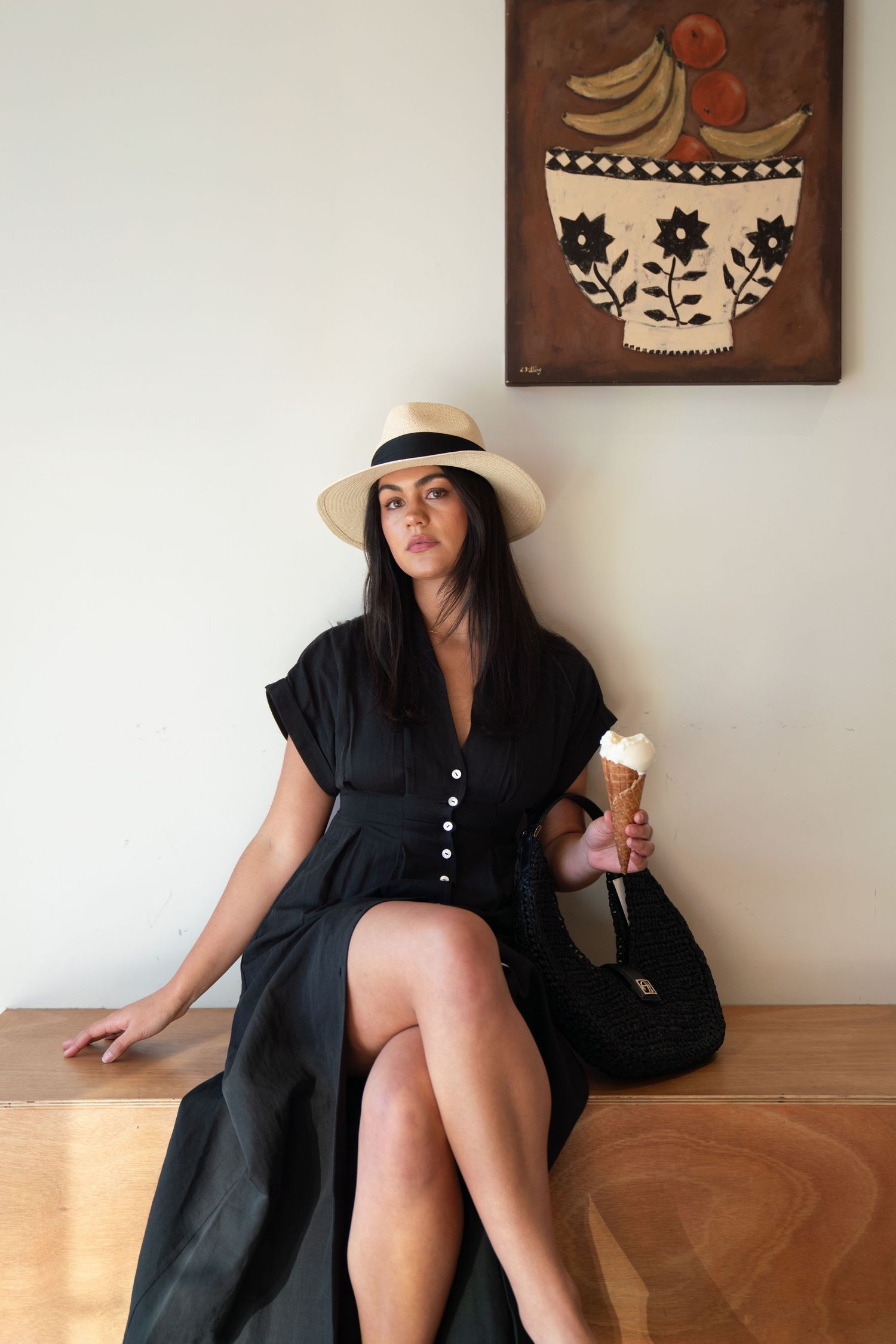Woman in black dress and straw hat holding ice cream cone, seated on bench near fruit bowl painting.