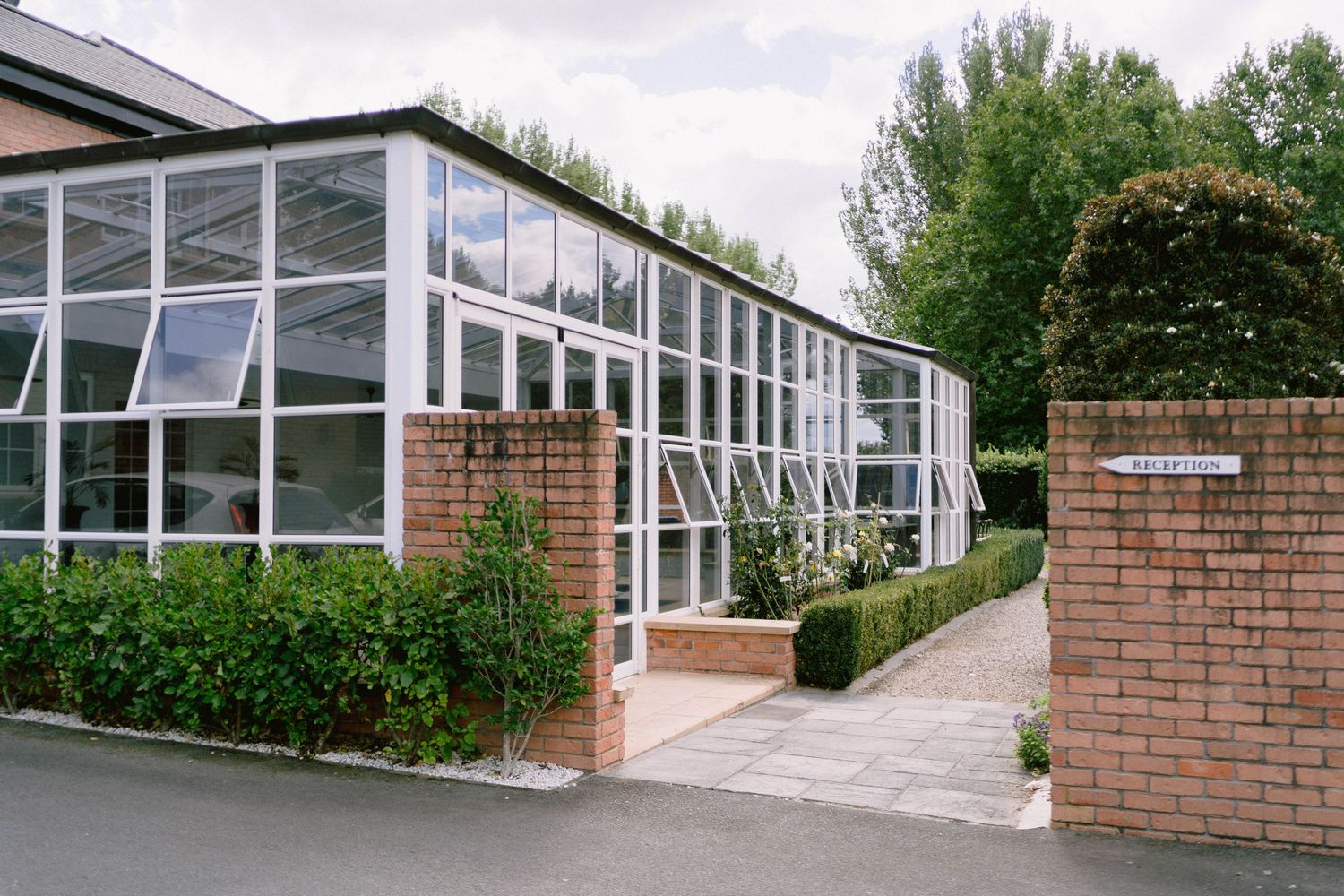 Modern glass and brick greenhouse structure with manicured hedges and walkway in an institutional setting.