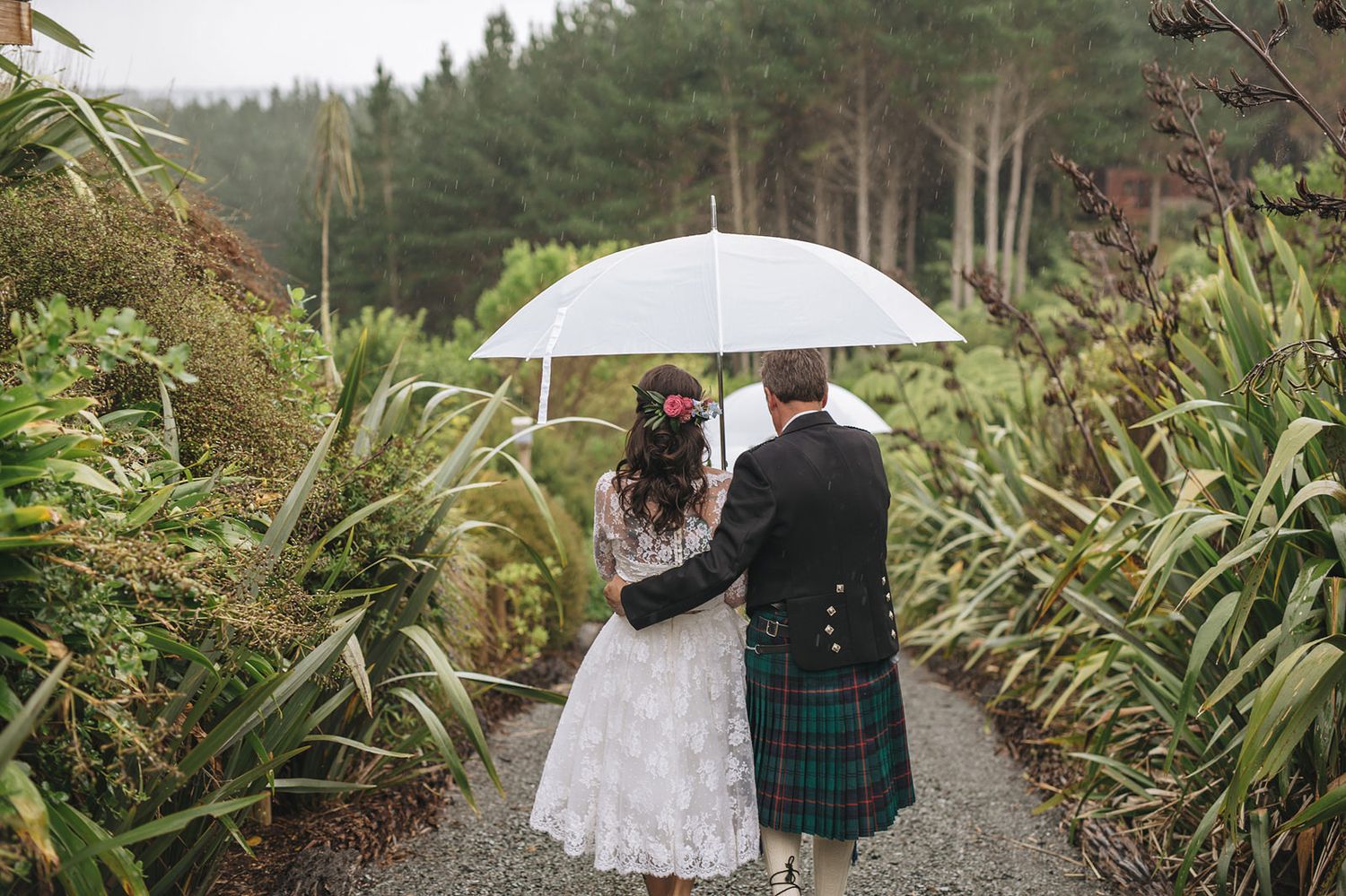 Wedding couple walks together on a forest path under white umbrella while surrounded by lush greenery and trees.