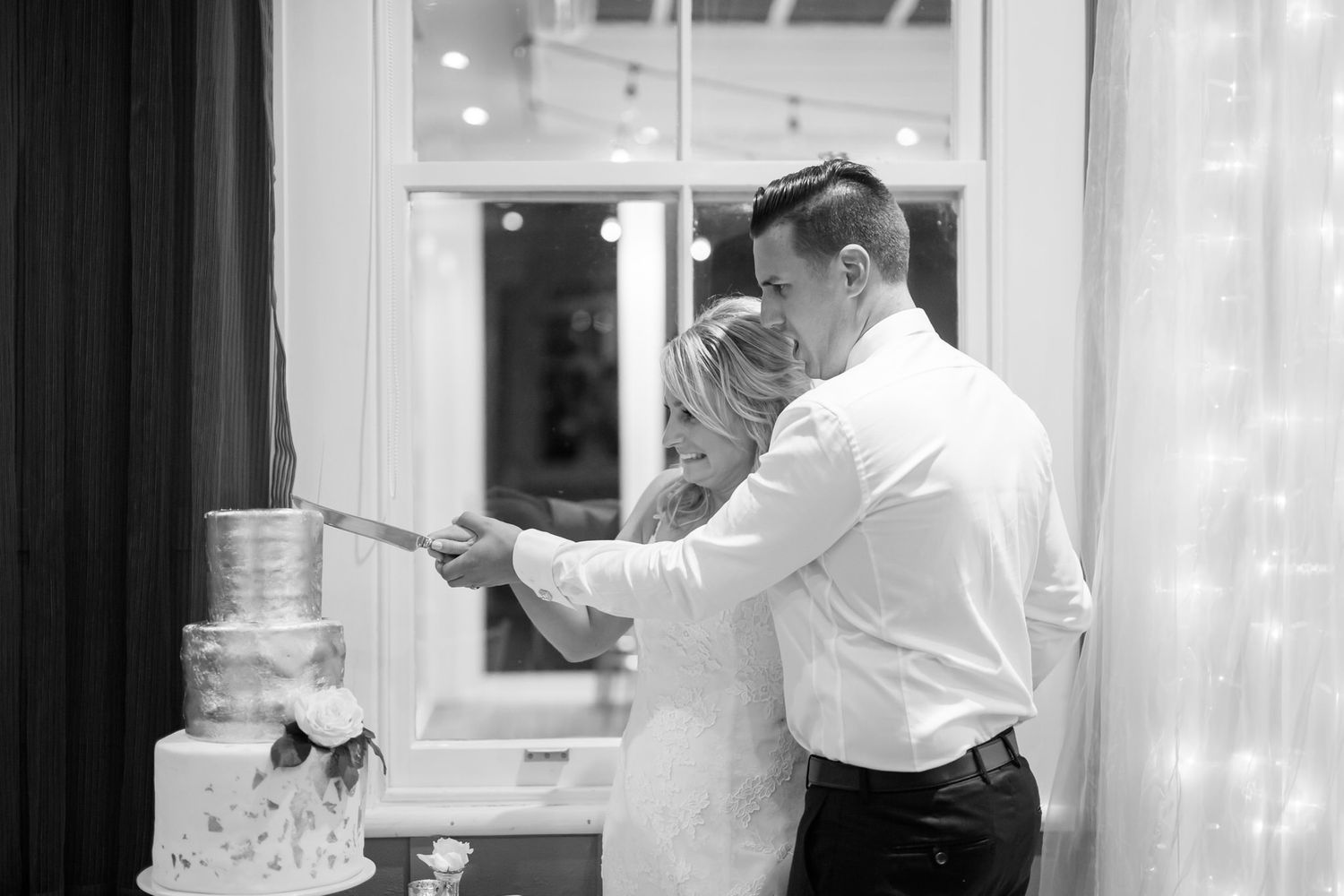 A couple in formal attire cuts their elegant white wedding cake together at their reception.