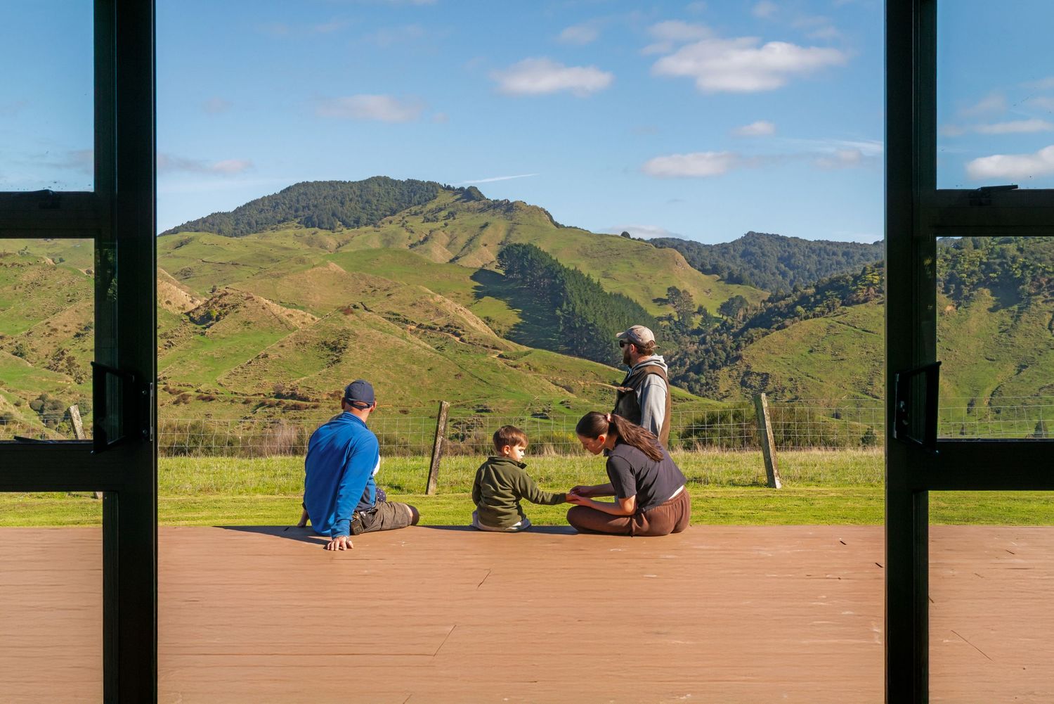 Group in casual attire sitting on wooden deck overlooking scenic green mountain landscape on sunny day.