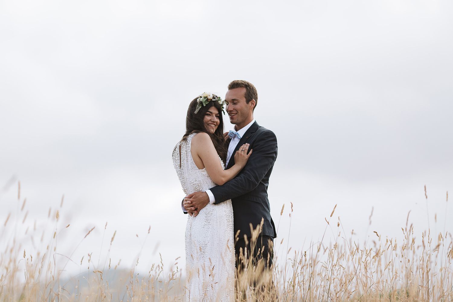 Newlywed couple embraces in romantic moment while standing in tall grass field on cloudy day.