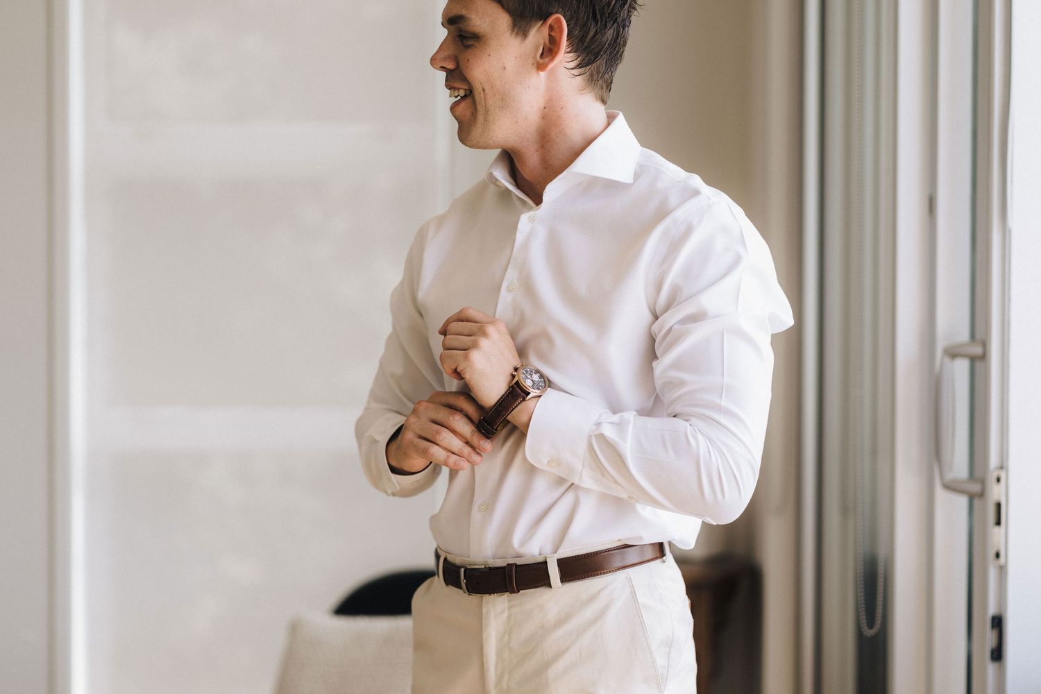 A person in a crisp white dress shirt and trousers smiles while adjusting their cuff by a bright window.