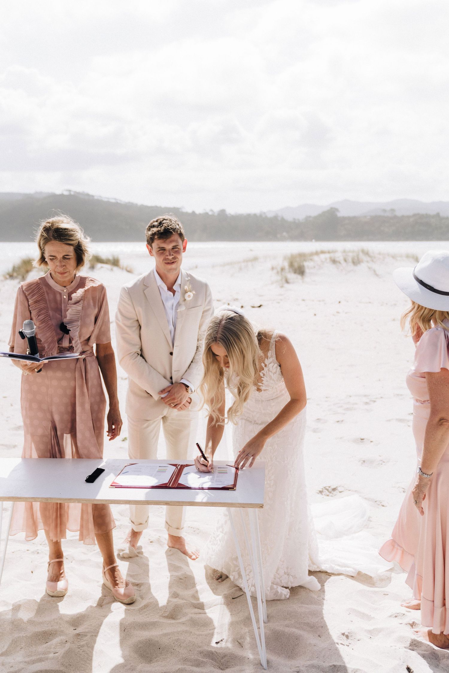 Wedding guests sign documents on a light wooden table at a beachside ceremony.