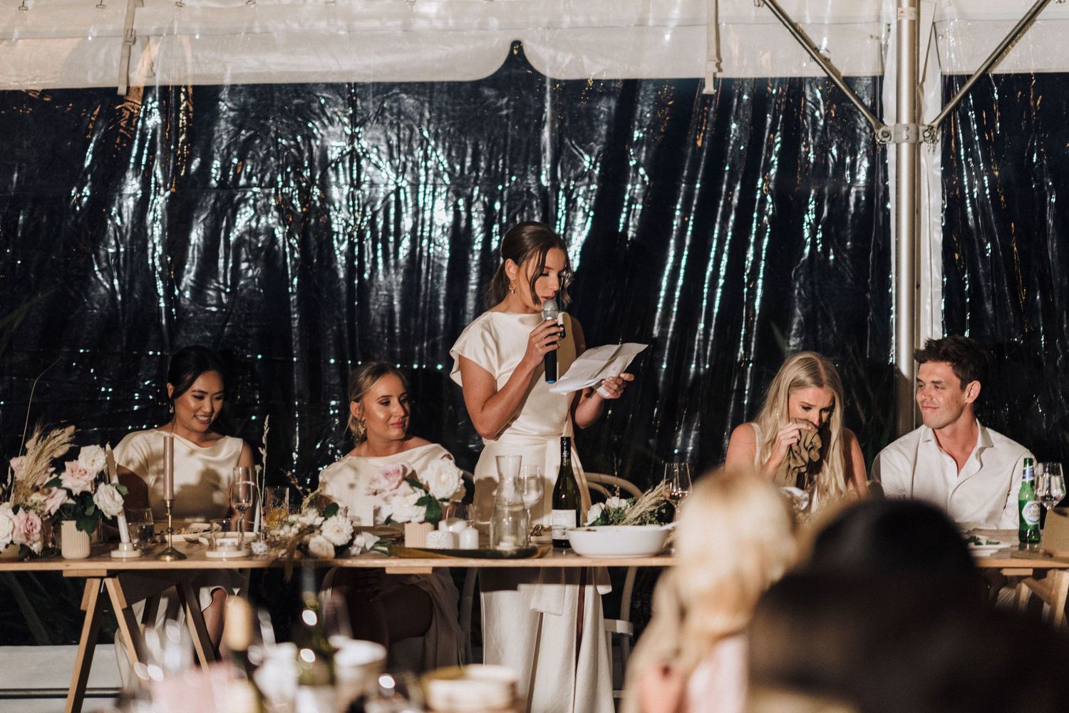 Wedding reception speeches being given at head table with black backdrop under tent.