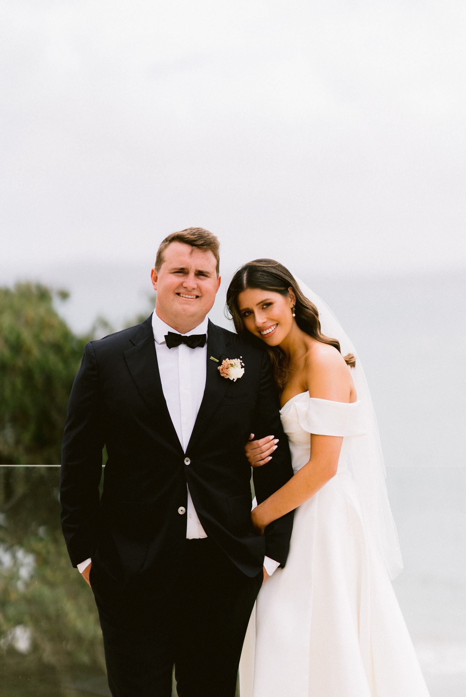 A bride and groom pose together outdoors on their wedding day, with the bride wearing an off-shoulder white dress.