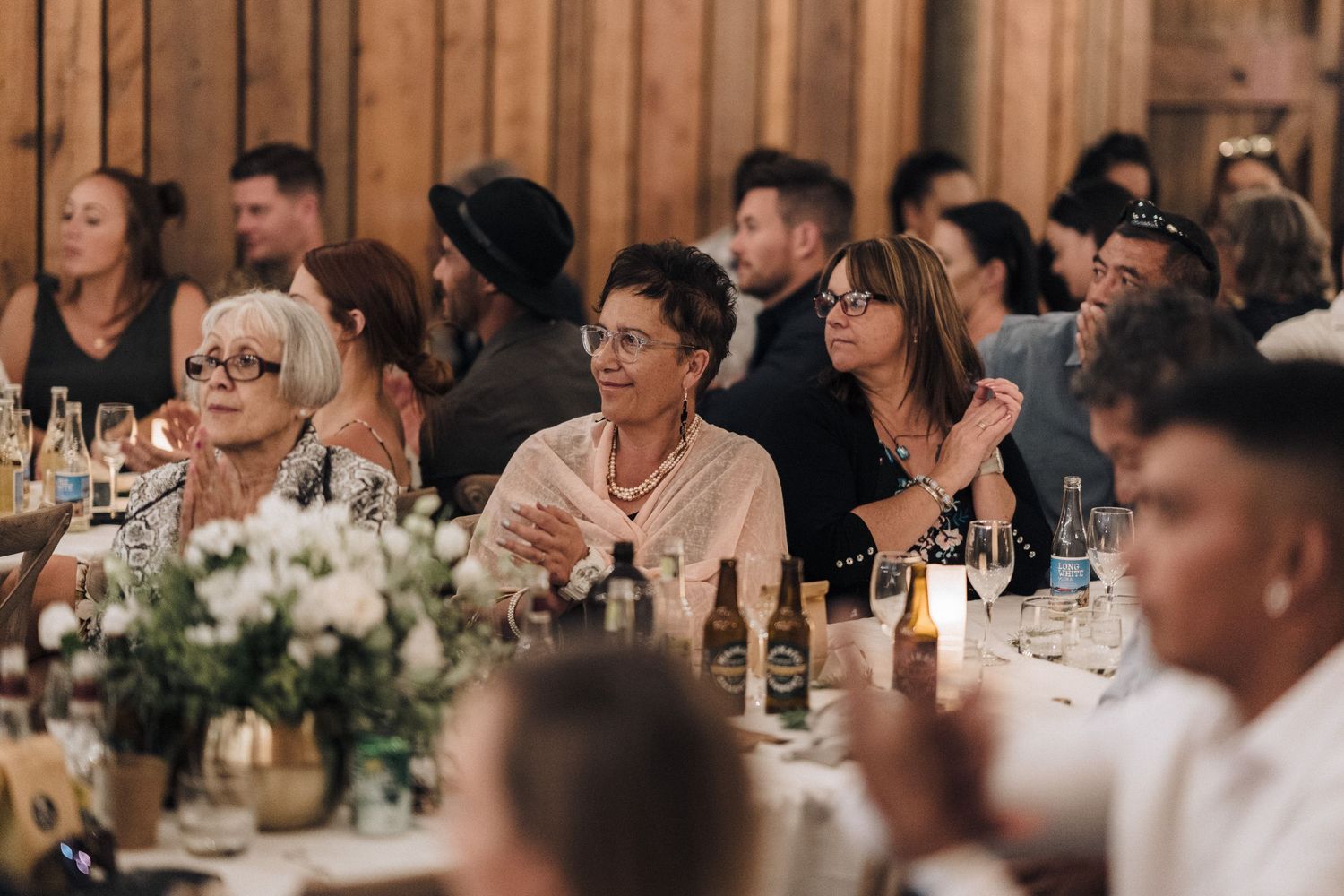 Wedding guests seated at long tables in a rustic wooden barn setting react with laughter and joy during celebrations.