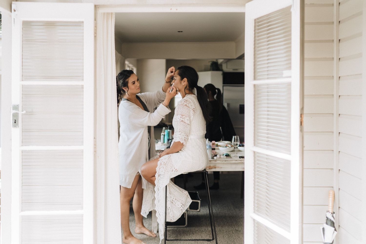 Two people in white robes share a tender makeup application moment in a bright, modern bathroom setting.