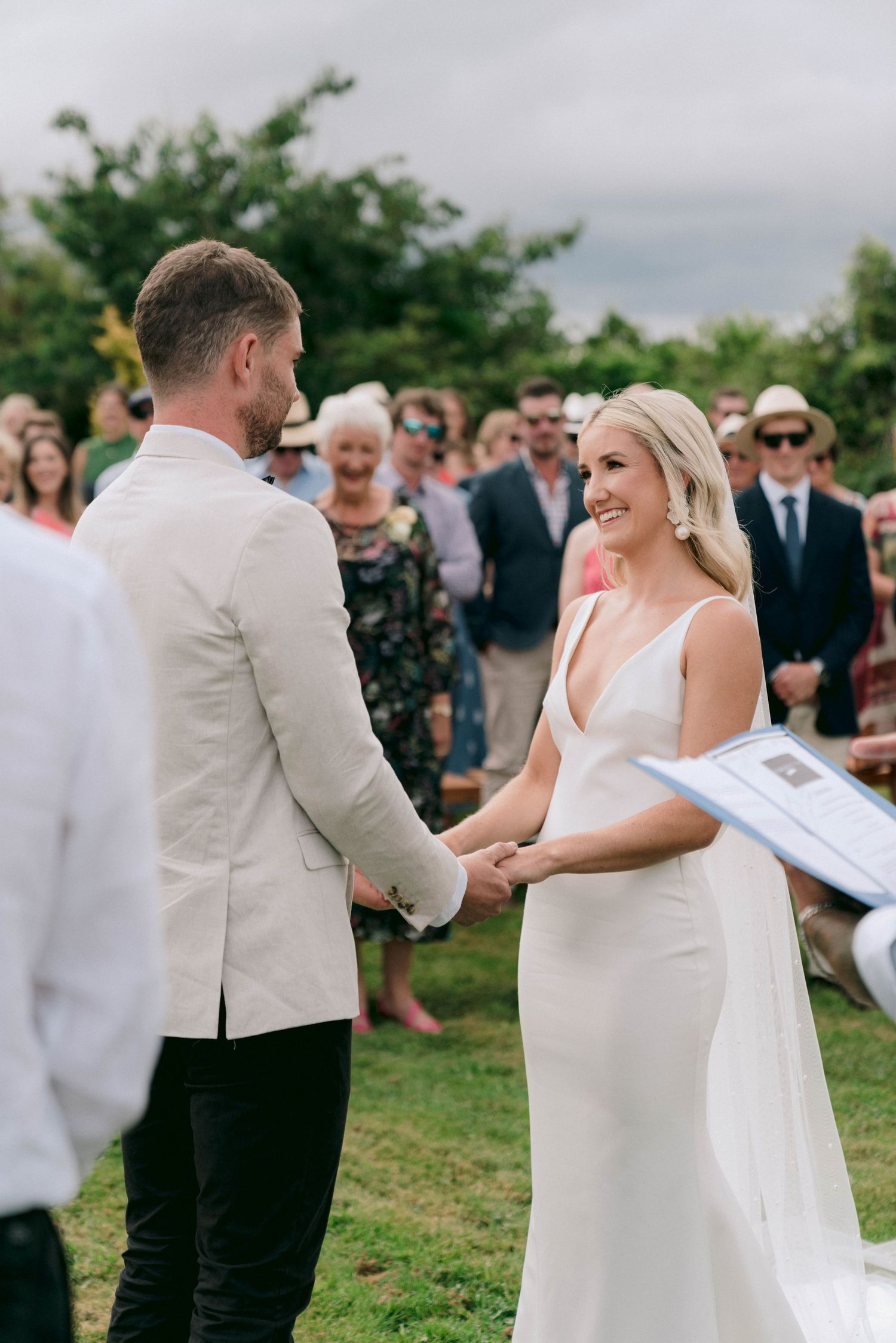 An intimate outdoor wedding ceremony shows a joyful exchange between a couple in white formalwear.