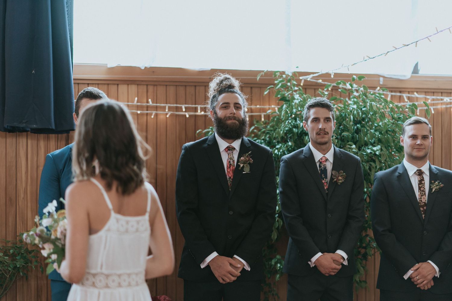 Wedding party stands formally arranged in a rustic wooden barn venue with string lights draped overhead.