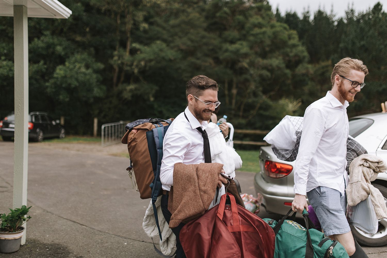 People in business attire gathering their luggage and belongings near parked cars in an outdoor area.