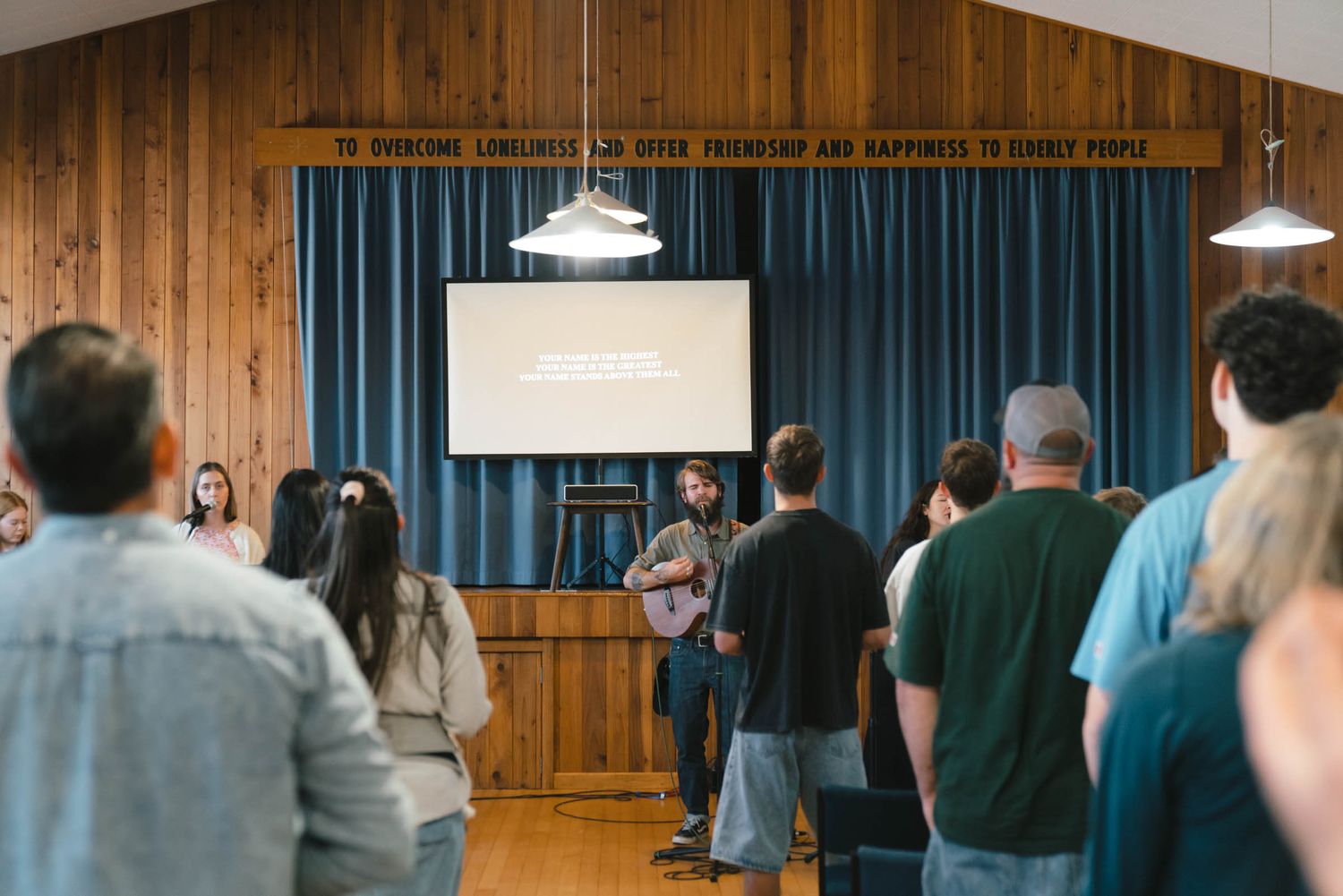 A congregation stands during worship in a wood-paneled church hall with blue curtains.