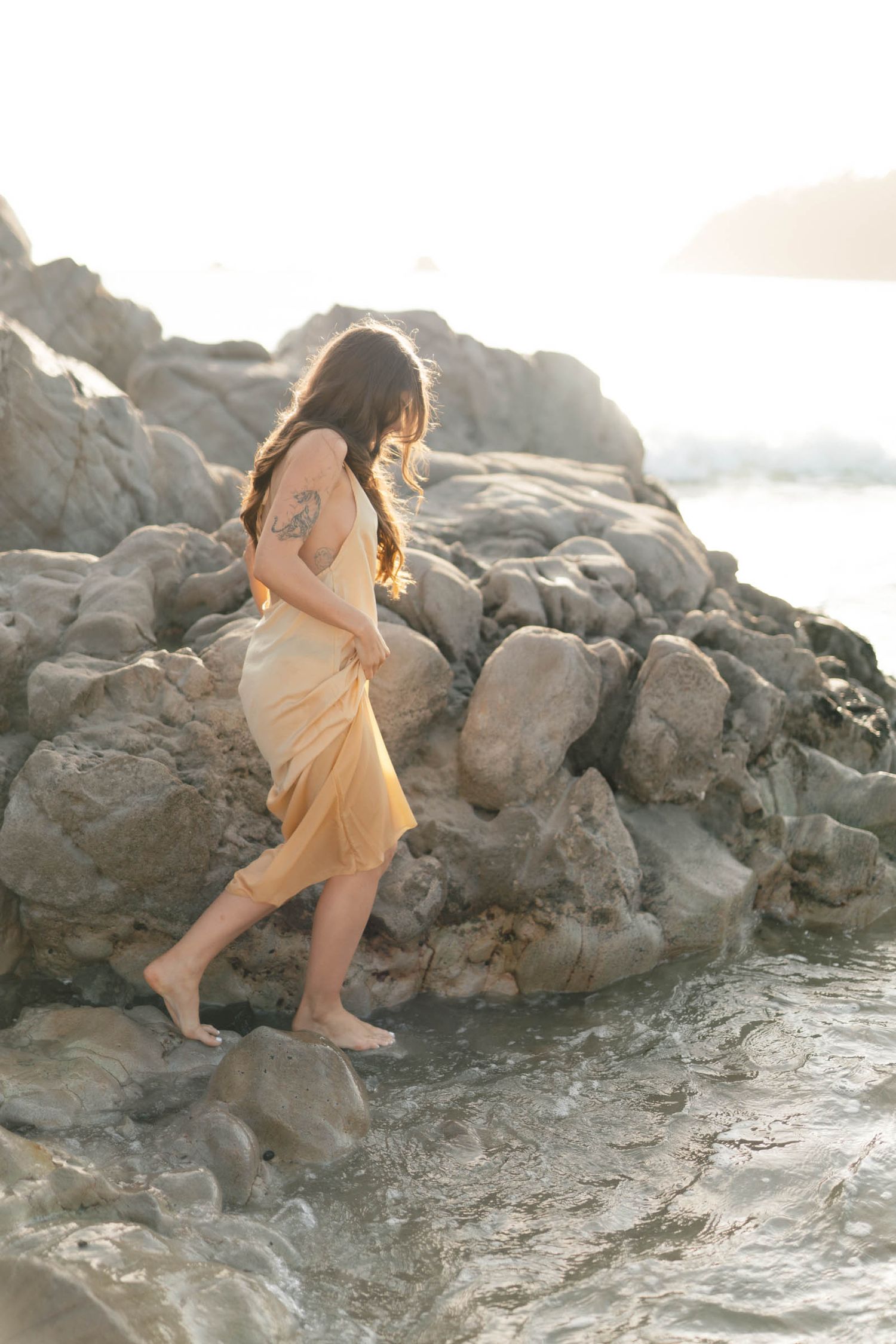 A person in a flowing dress carefully traverses rocky coastal terrain at golden hour.