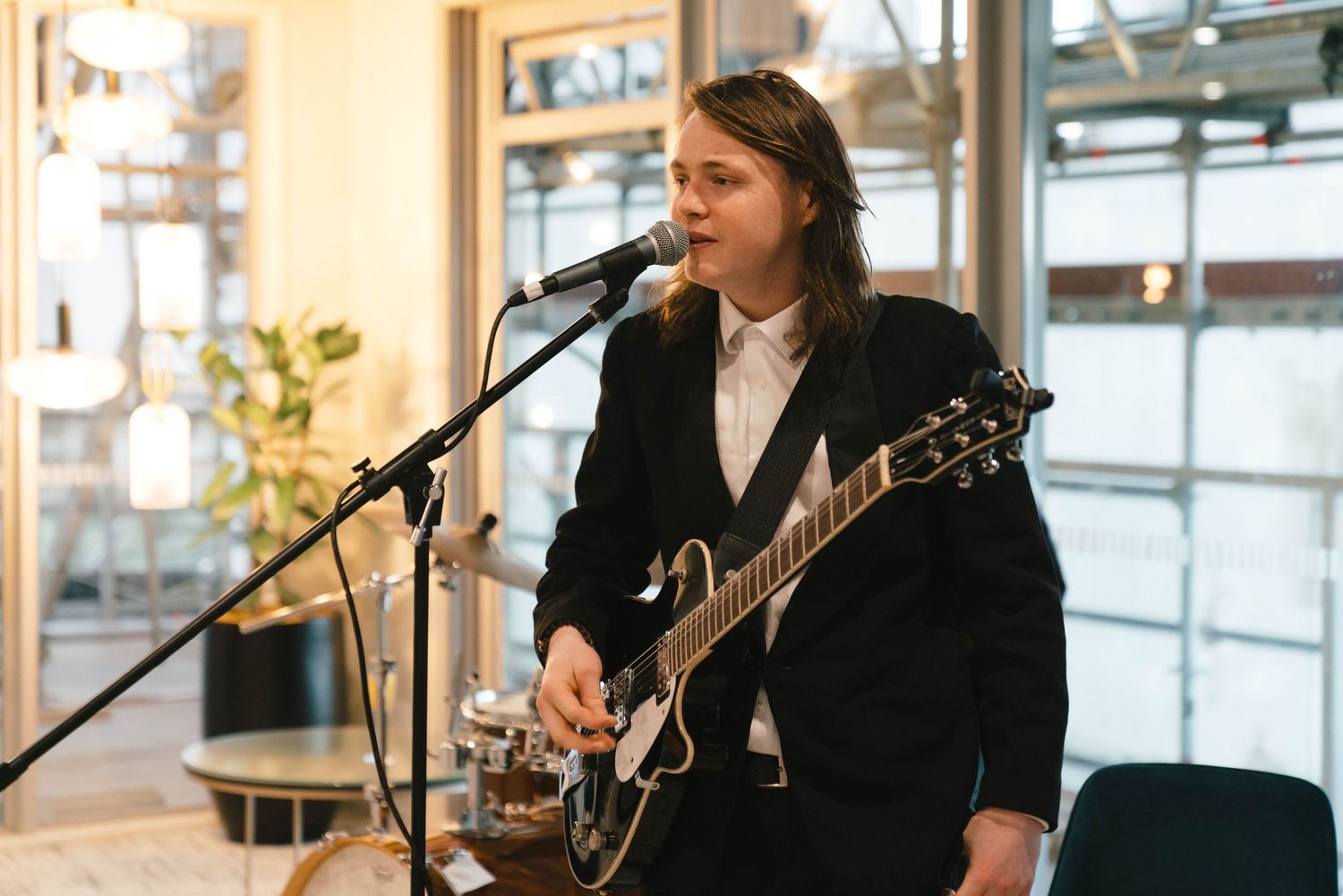 Musician in black jacket plays electric guitar while singing into microphone at indoor venue near windows.