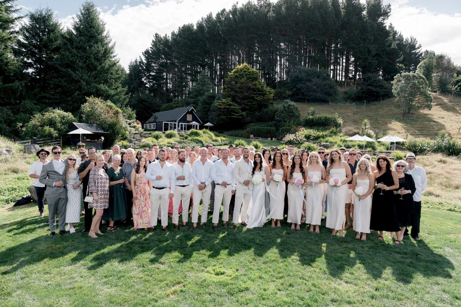 Wedding party members lined up for a group photo on a hillside with evergreen trees in background.